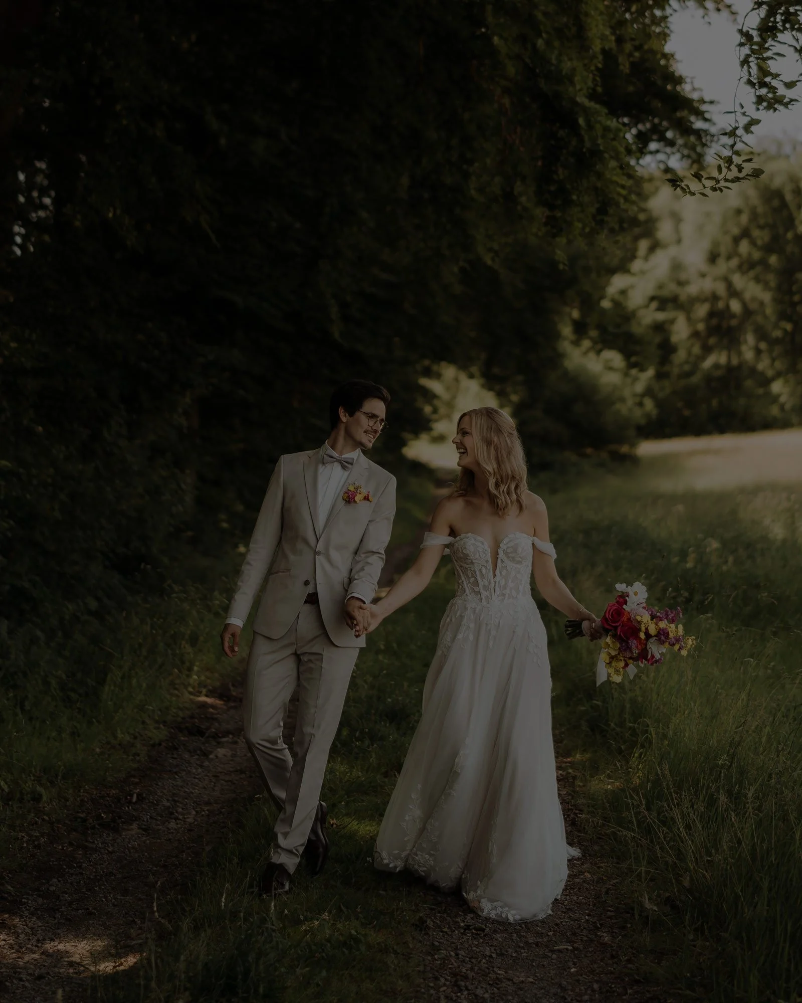 Ein Brautpaar in Hochzeitskleidung beim Spaziergang im Freien. Sie sind von goldenem Licht und dunkelgrünem Wald umgeben.