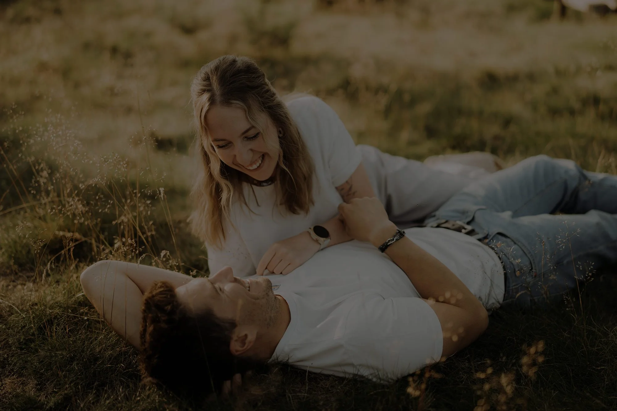 Ein lächelndes Paar liegt auf der Wiese im Freien und blickt sich liebevoll an.