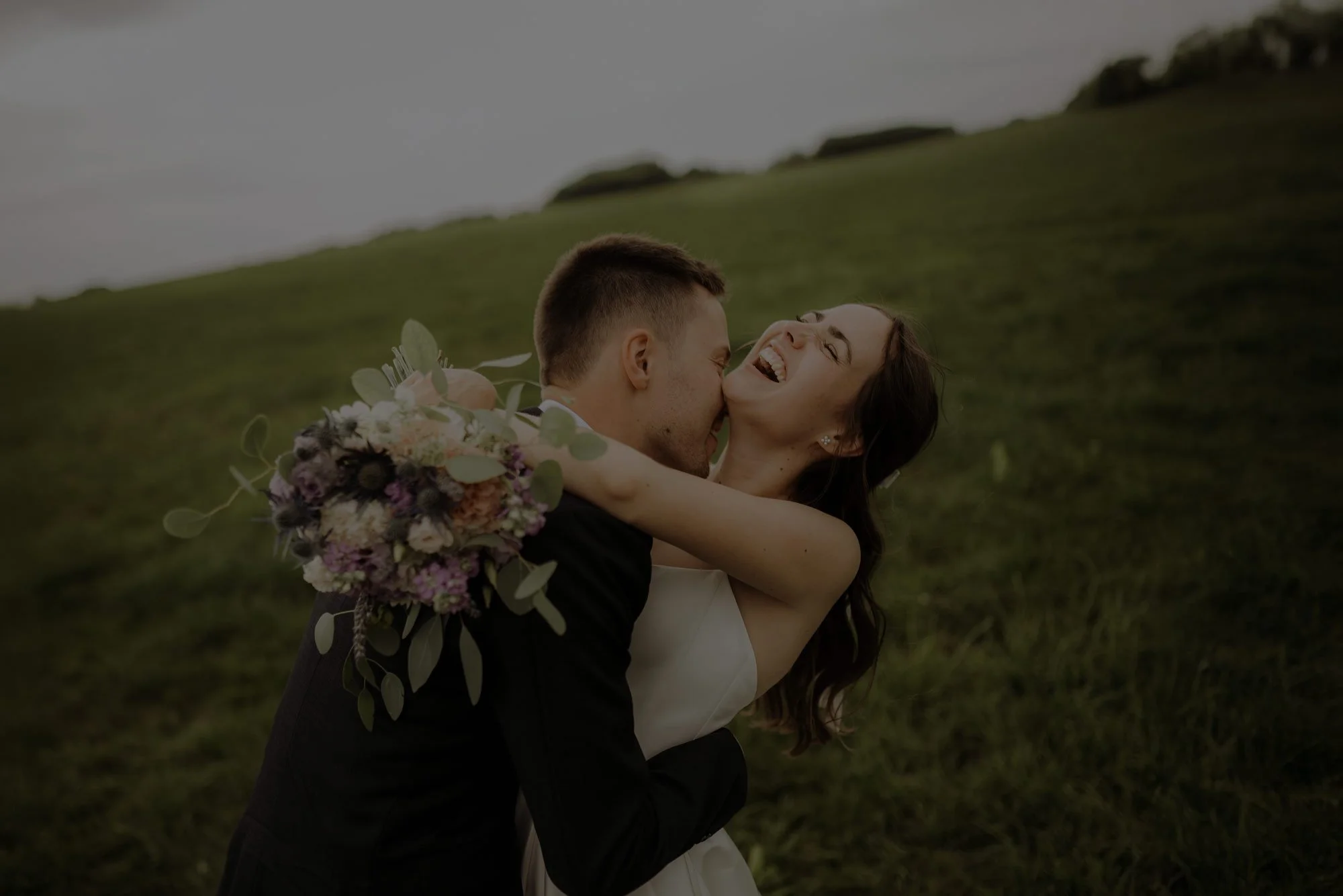 Ein lachendes Paar, das sich umarmt, in einem grünen Feld bei Sonnenuntergang. Der Mann küsst der Frau den Hals hinunter.