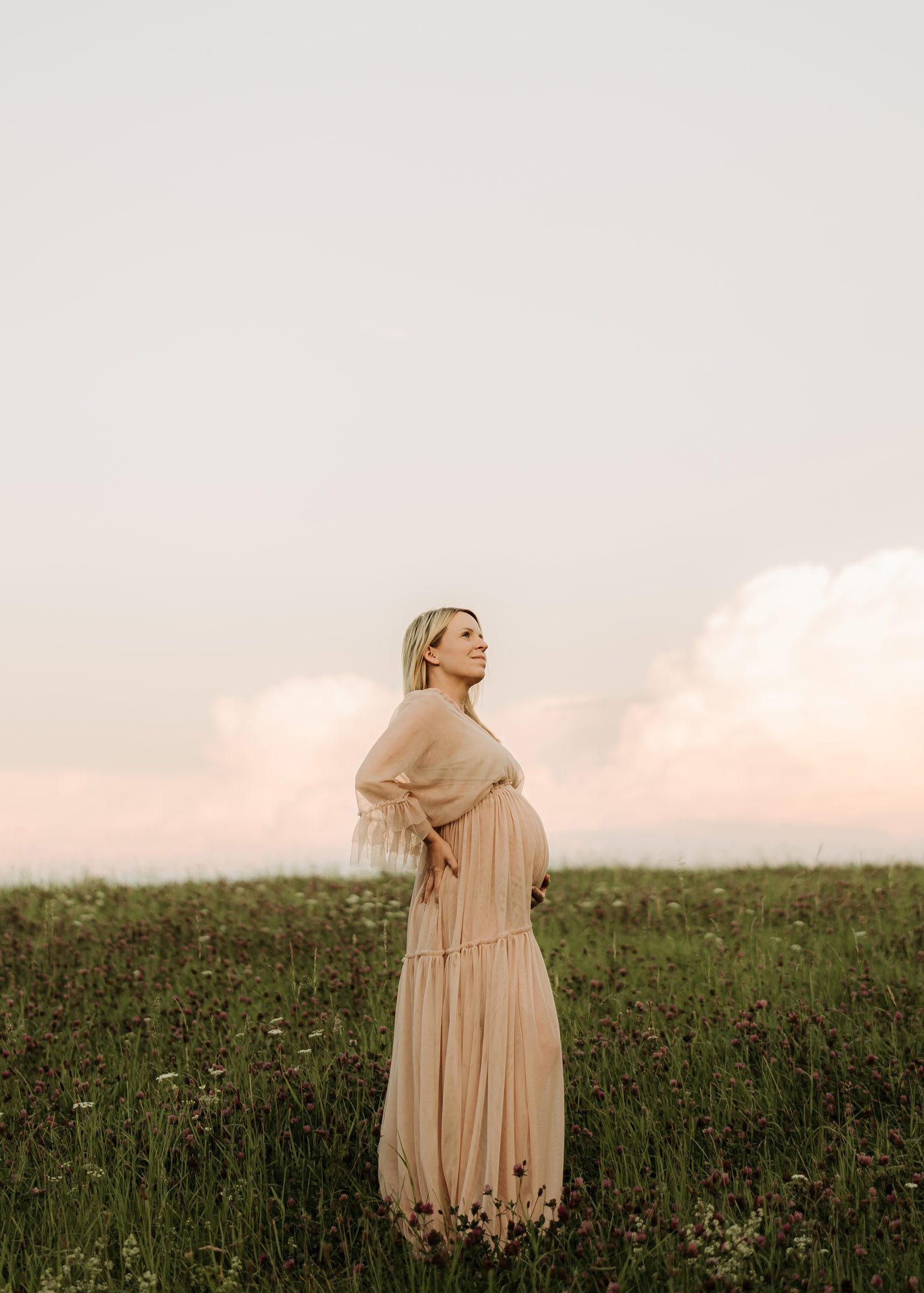 Schwangere Frau in einem langem, beigen Kleid steht in einer grünen Blumenwiese bei Sonnenuntergang und blickt nach oben.