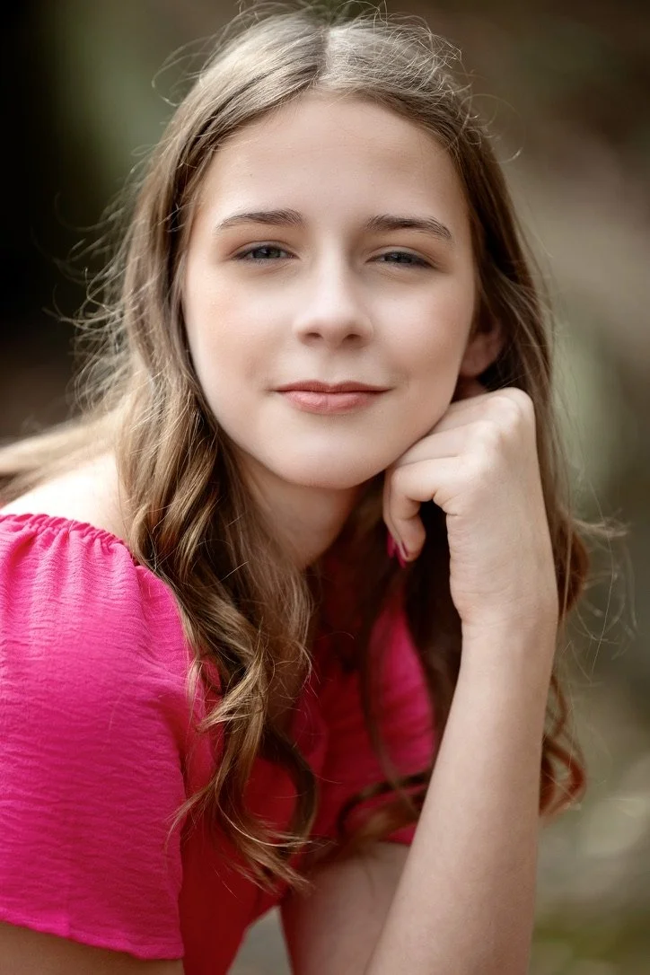 A young woman with long wavy blonde hair and a fair complexion, wearing a pink top, resting her chin on her hand and looking at the camera with a gentle smile outdoors.
