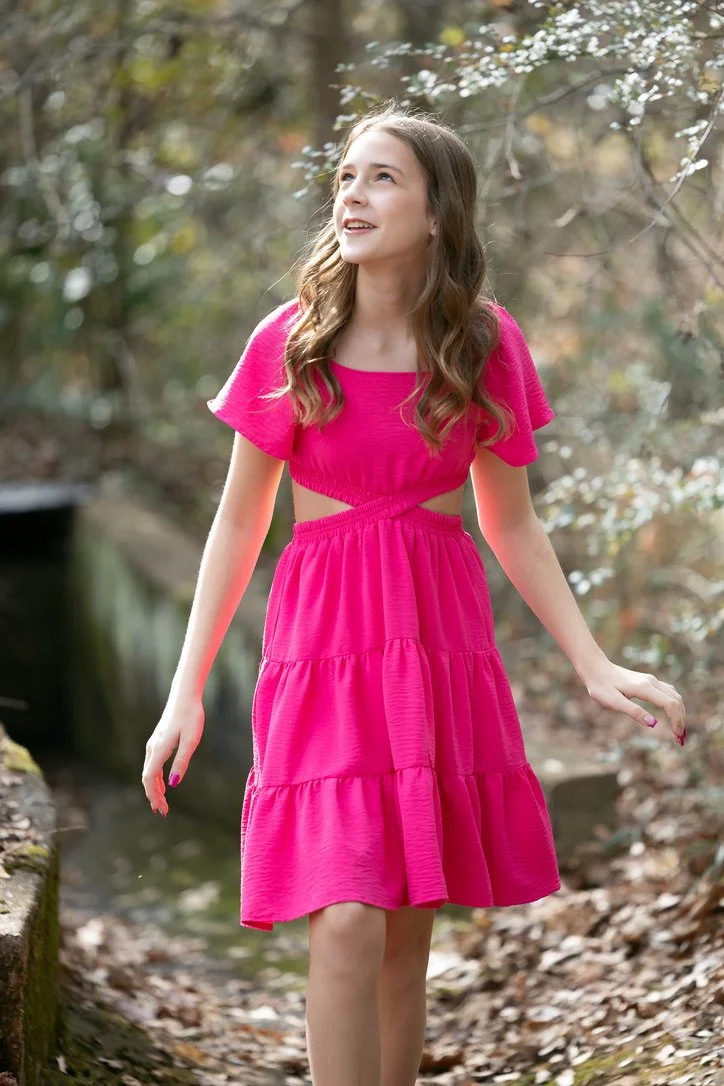 A young girl with long brown hair wearing a bright pink dress and walking outdoors through a wooded area.