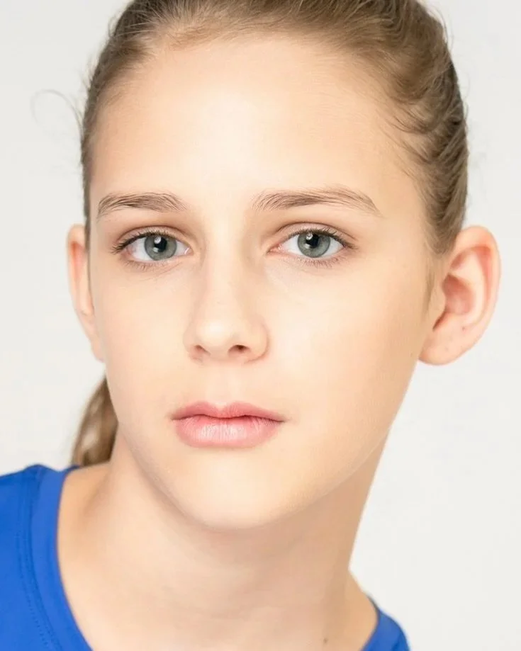 A young girl with blue eyes, blonde hair tied back, wearing a blue shirt, looking directly at the camera with a neutral expression.