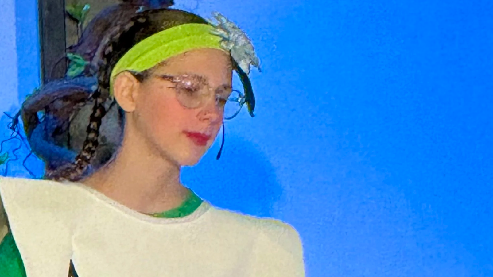 Person dressed as an ace of spades playing card standing on stage, wearing a costume that resembles a large playing card, with green leggings and a flower headband.