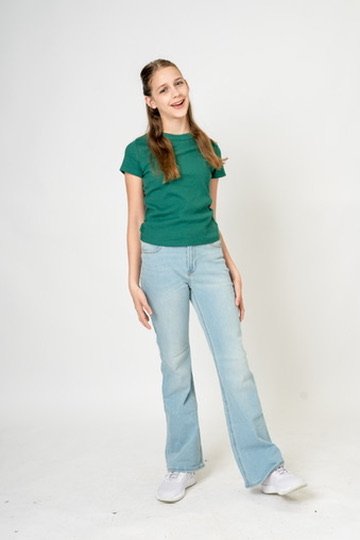 A young woman with long brown hair wearing a green t-shirt, light blue jeans, and white sneakers, standing against a plain white background, smiling with her hands relaxed at her sides.