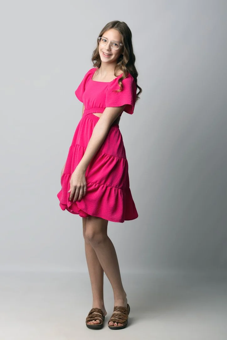 Young woman with glasses in a pink dress and brown sandals standing against a gray background.