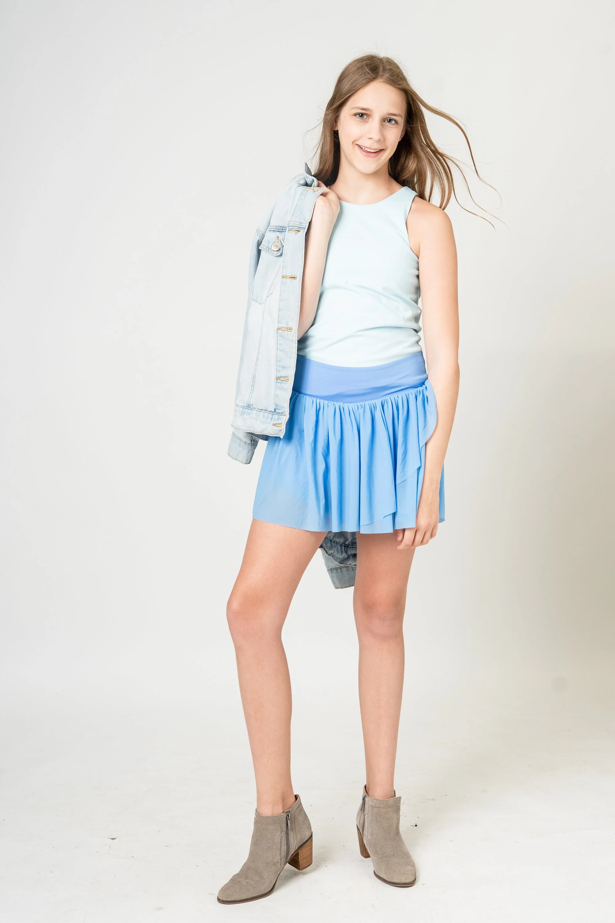 A young woman with long hair, smiling, wearing a sleeveless light blue top, a darker blue skirt, gray ankle boots, and carrying a denim jacket over her shoulder, standing against a plain white background.