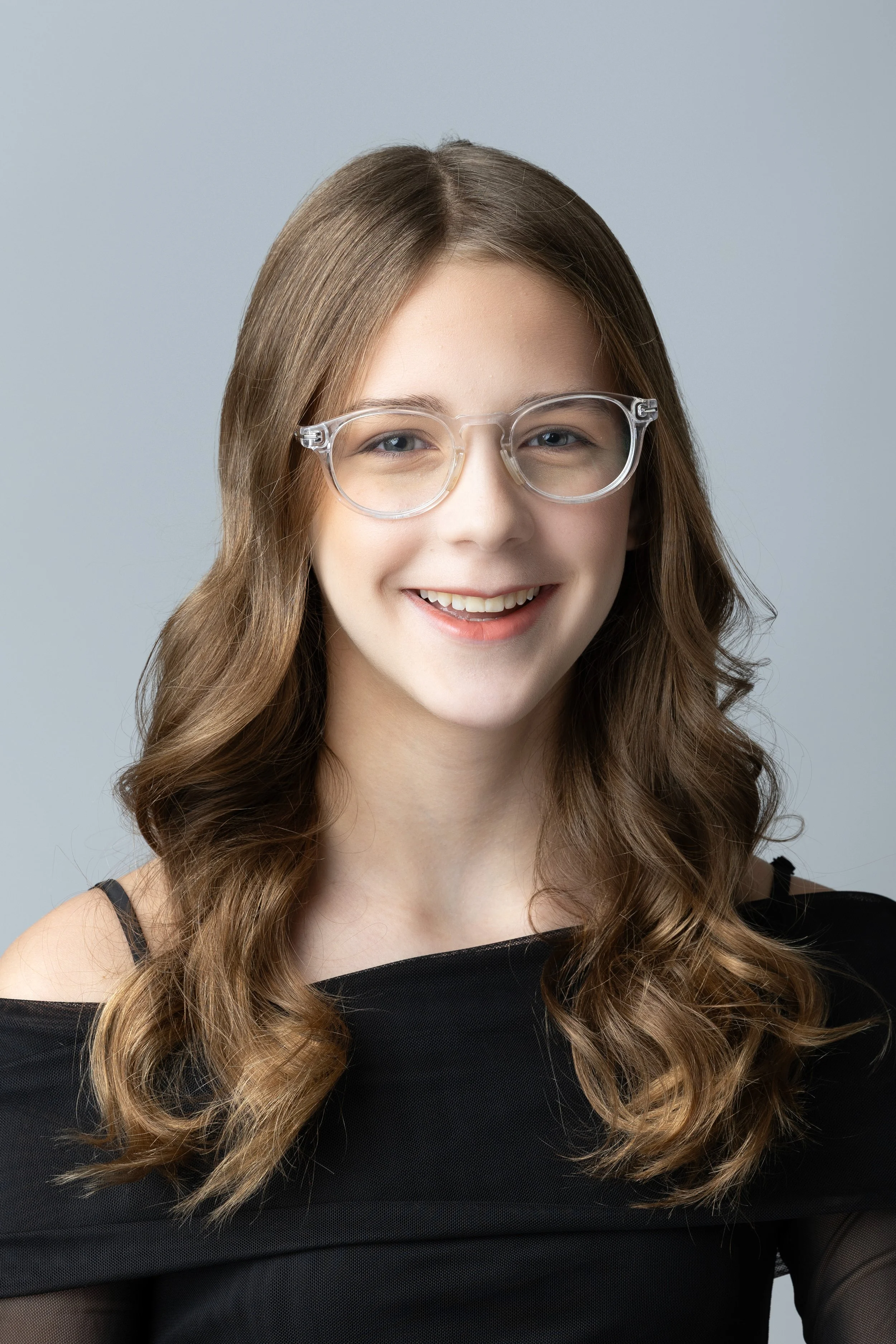 A young woman with long wavy brown hair wearing clear glasses and a black off-shoulder top, smiling at the camera against a plain gray background.