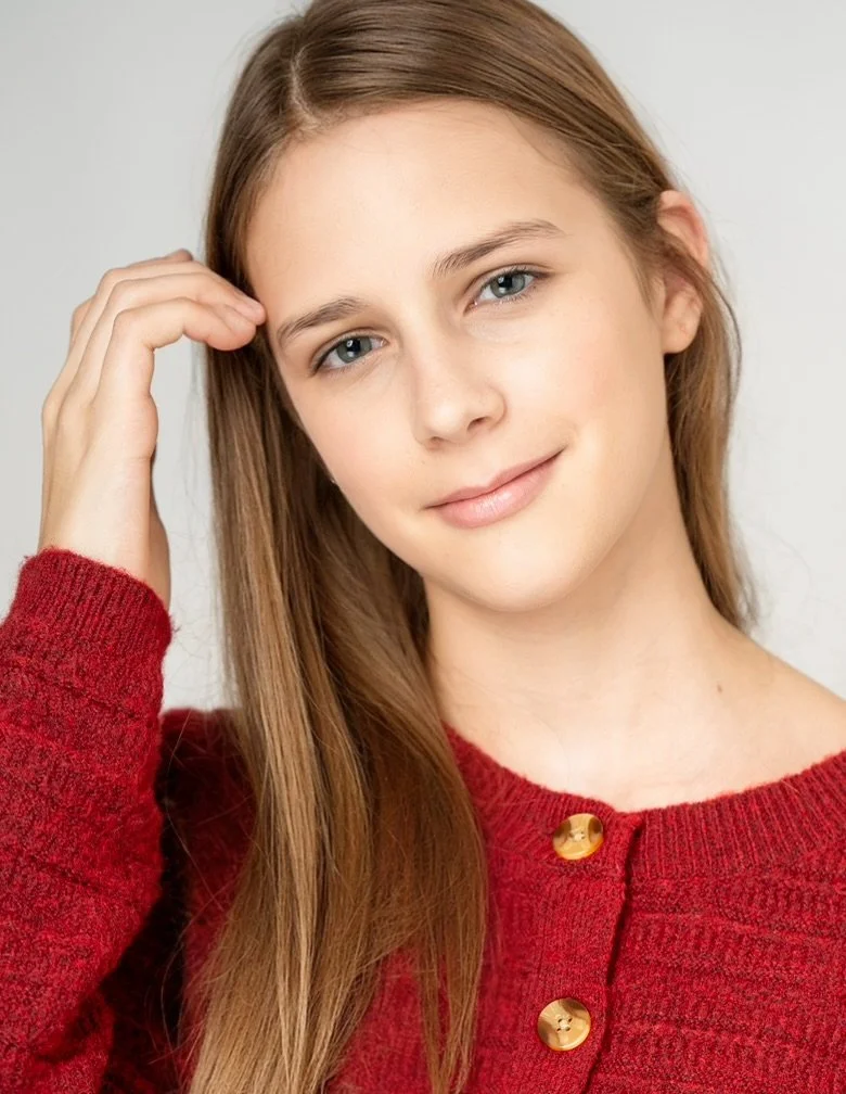 A young woman with long brown hair and blue eyes, wearing a red knit sweater with gold buttons, smiling softly, touching her head with her right hand.