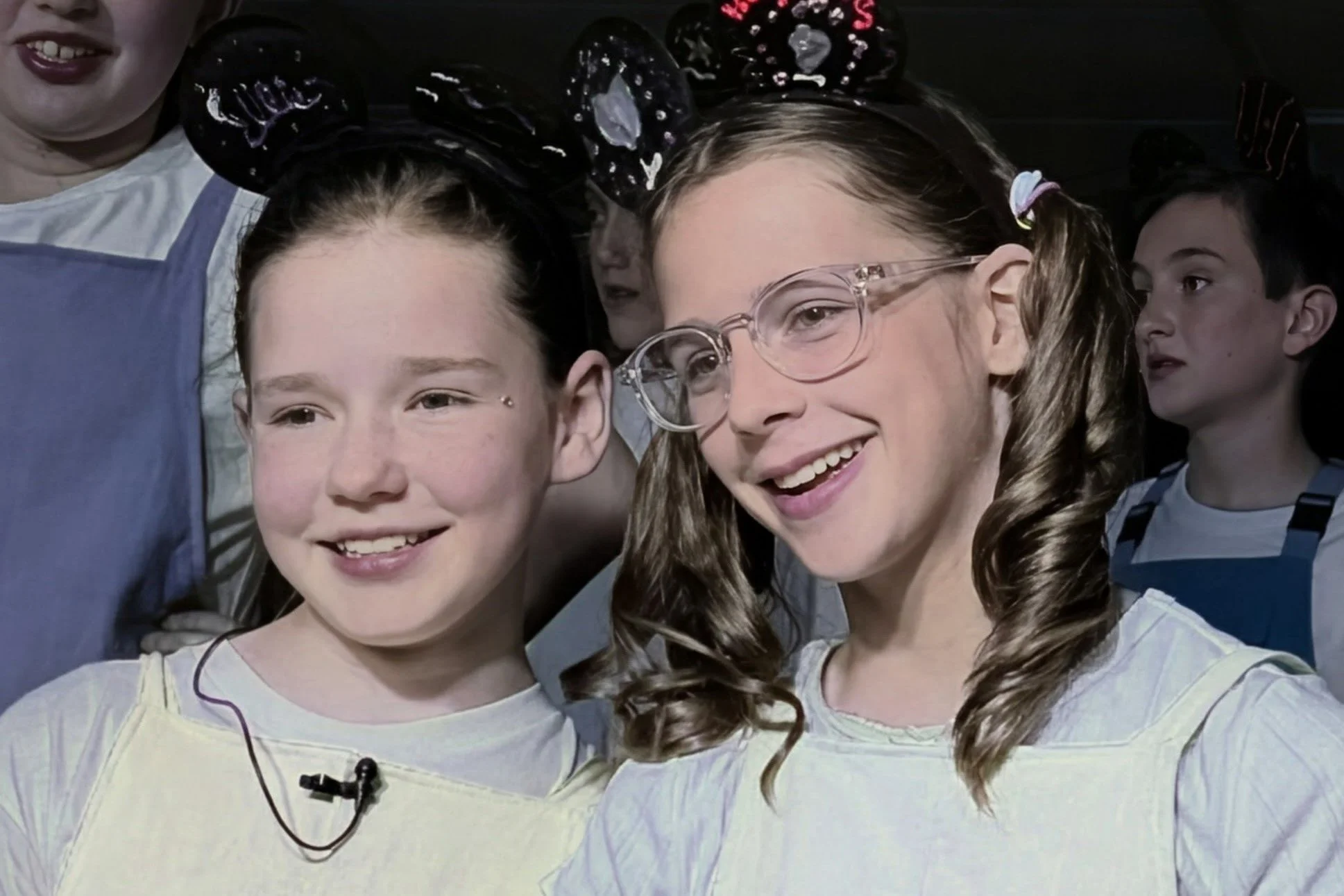Two women smiling and posing with their arms around each other, wearing Mickey Mouse ear hats with polka dot patterns. One woman has curly hair, glasses, and pigtails, while the other has straight hair. The photo appears to be taken indoors in black and white.