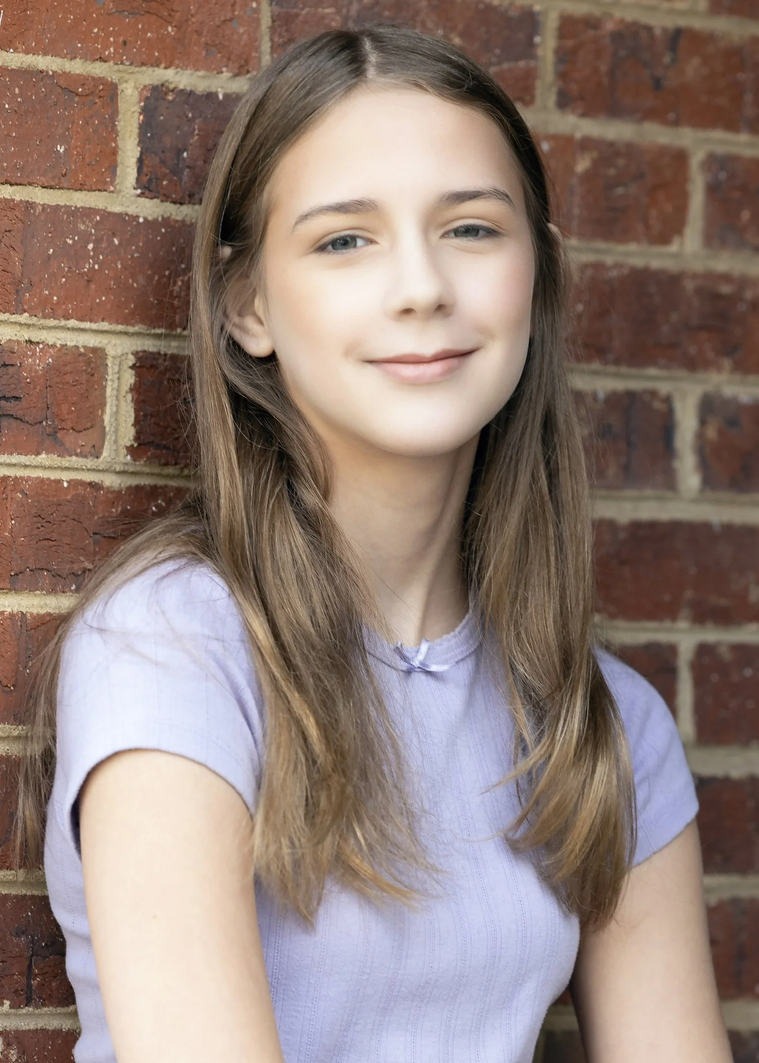 A young girl with long brown hair and a slight smile, wearing a light purple t-shirt, standing against a red brick wall.