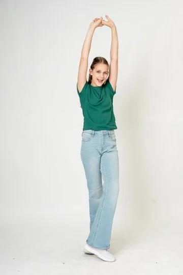 A young woman in a green t-shirt and light blue jeans stretching with arms raised above her head.