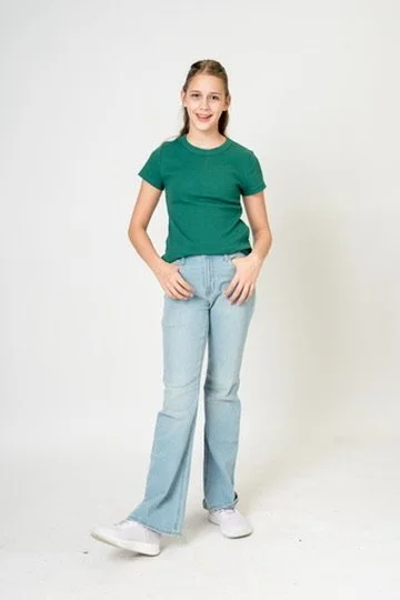 A young girl with long hair, wearing a green t-shirt and light blue jeans, standing against a plain white background.