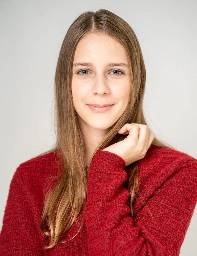 A young woman with long, light brown hair and blue eyes, smiling softly, wearing a red sweater, posing against a plain light gray background.