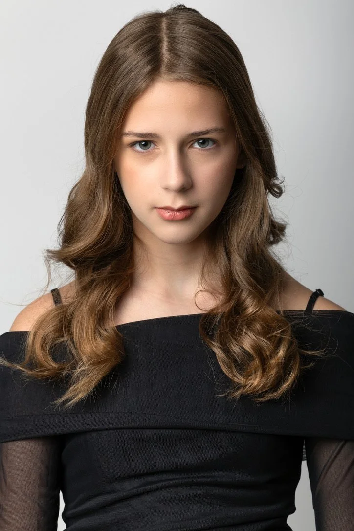 A young woman with long, wavy brown hair wearing a black off-the-shoulder top against a plain gray background.