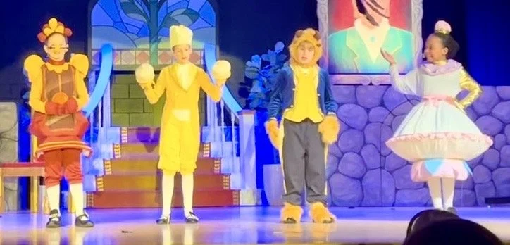 Young girl dressed as a chef, wearing a white chef's hat, yellow jacket, and holding two white oven mitts, standing indoors with a neutral background.