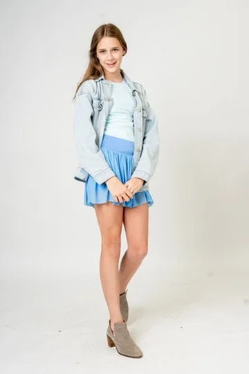 A young girl with long hair wearing a light blue t-shirt, a denim jacket, a blue skirt, and tan ankle boots standing against a plain white background.