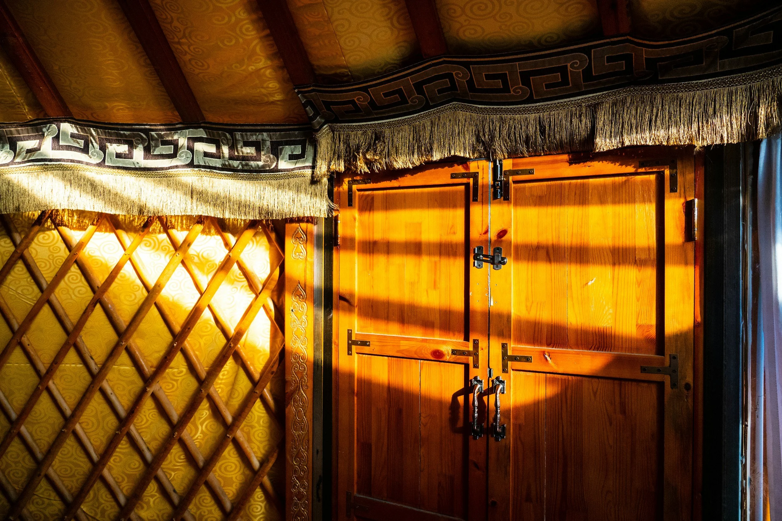 Sunlight shining on a wooden door with black metal hinges and locks, beside a yellow wall with a lattice pattern. Clothing with fringes and patterned border hanging above the door.
