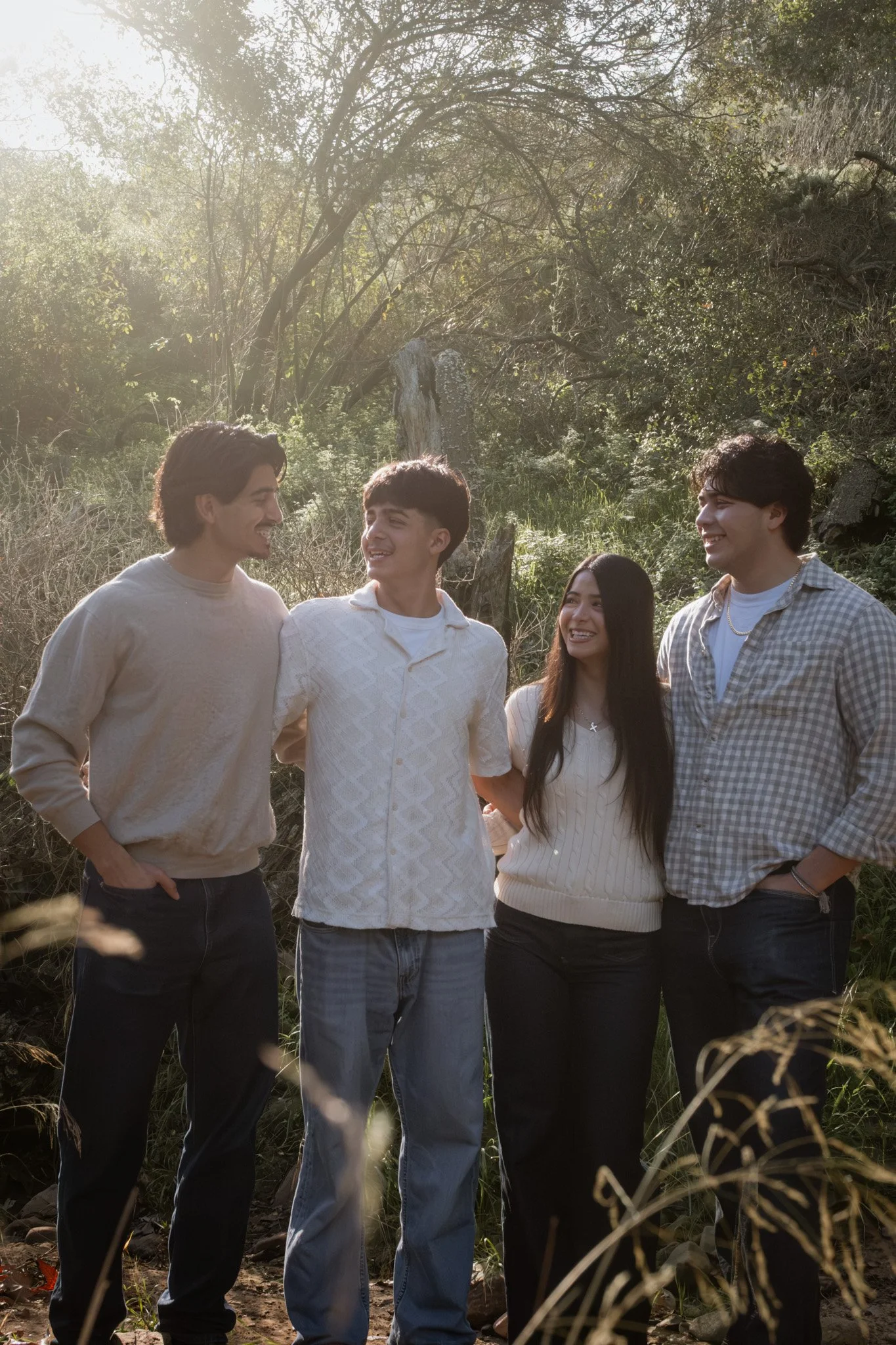 Four young friends standing outdoors in a sunlit natural setting, smiling and looking at each other.