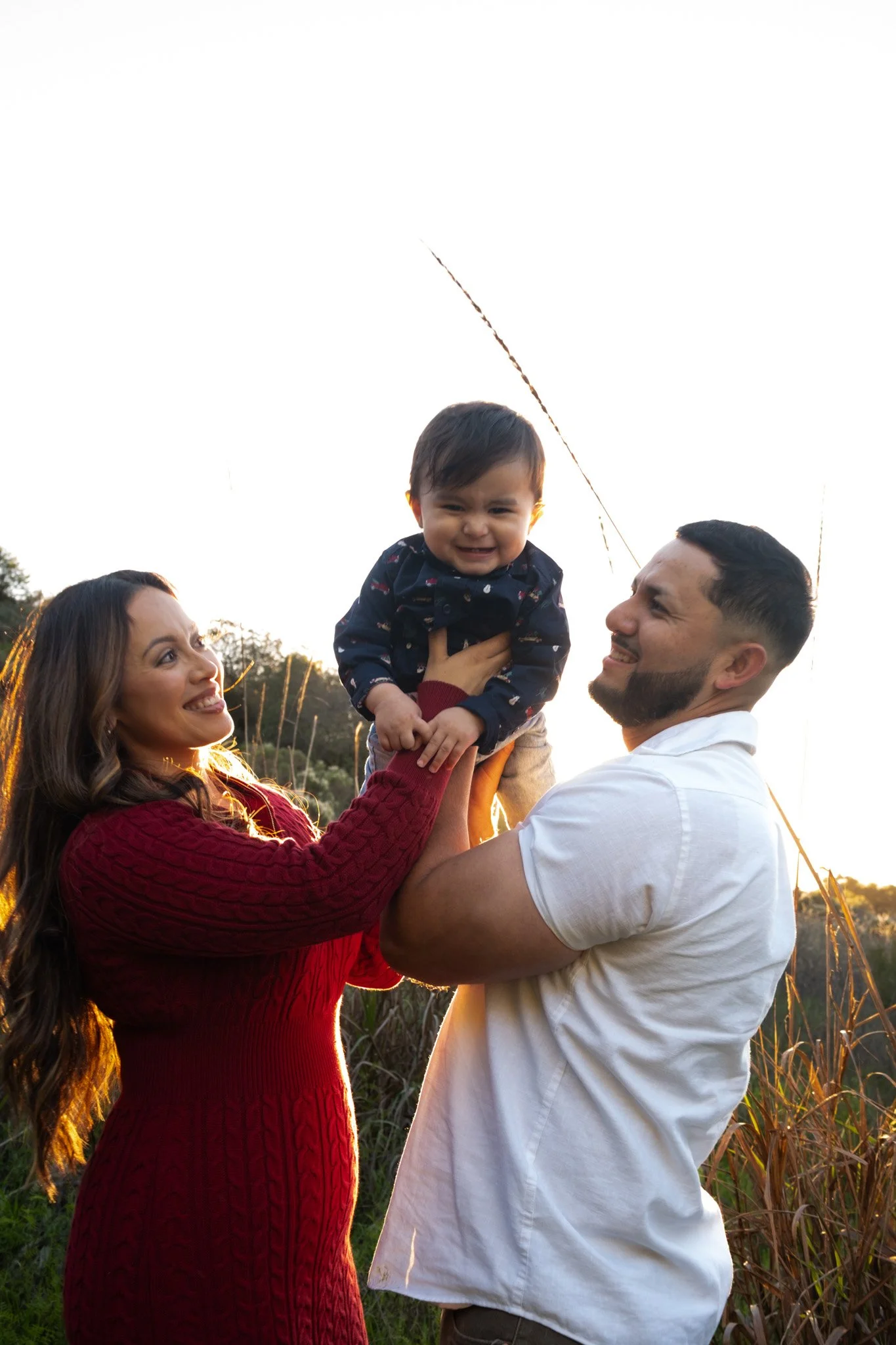 A family of three outdoors at sunset, with a woman in a red sweater holding a smiling toddler, and a man in a white shirt supporting the toddler.