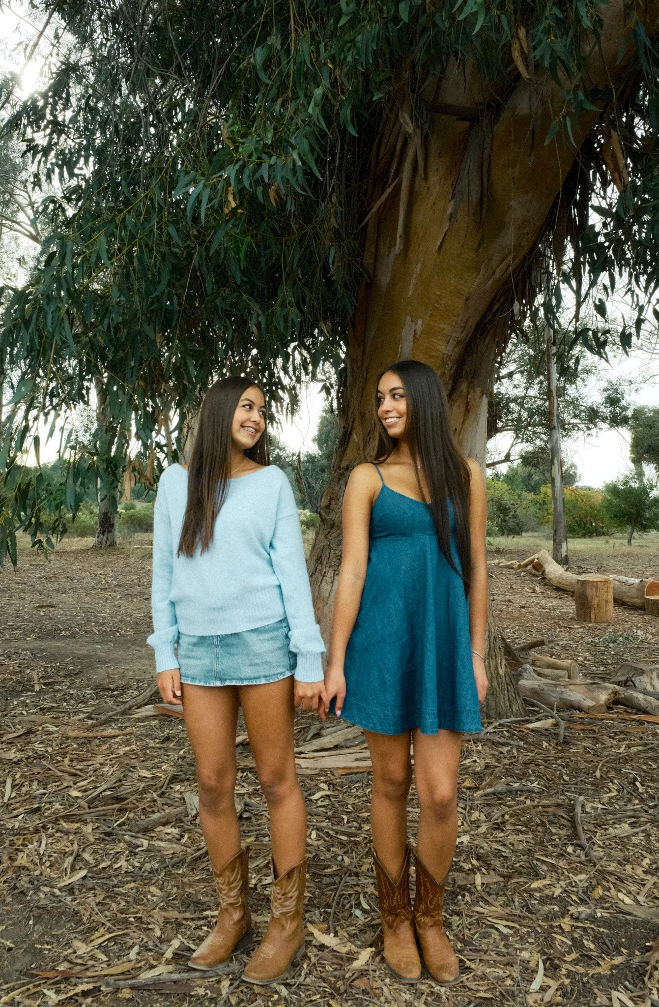 Two women standing outdoors near a large tree, holding hands and smiling at each other.