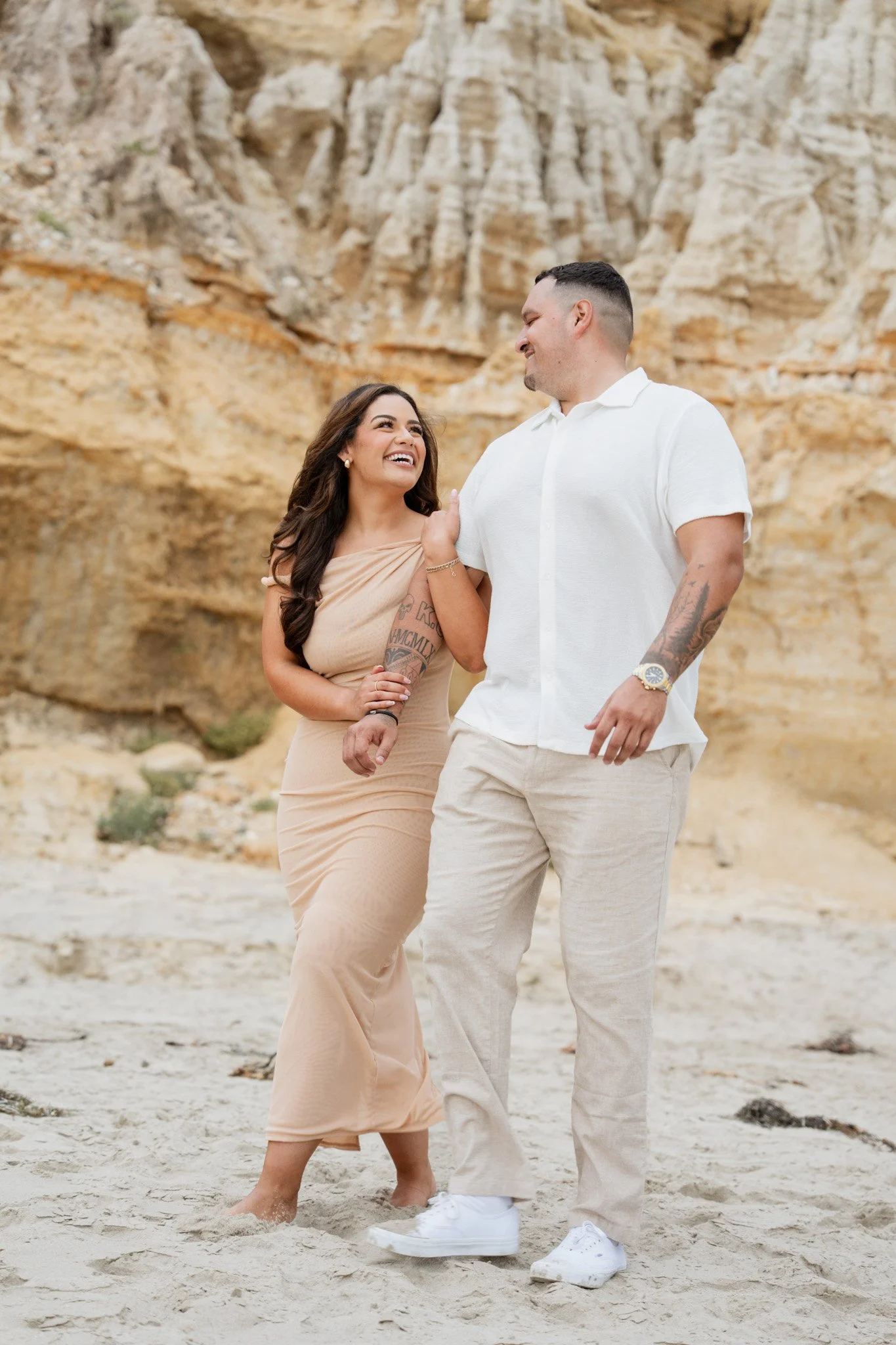 A smiling woman and man walking on a sandy beach with cliffs in the background, enjoying each other's company.