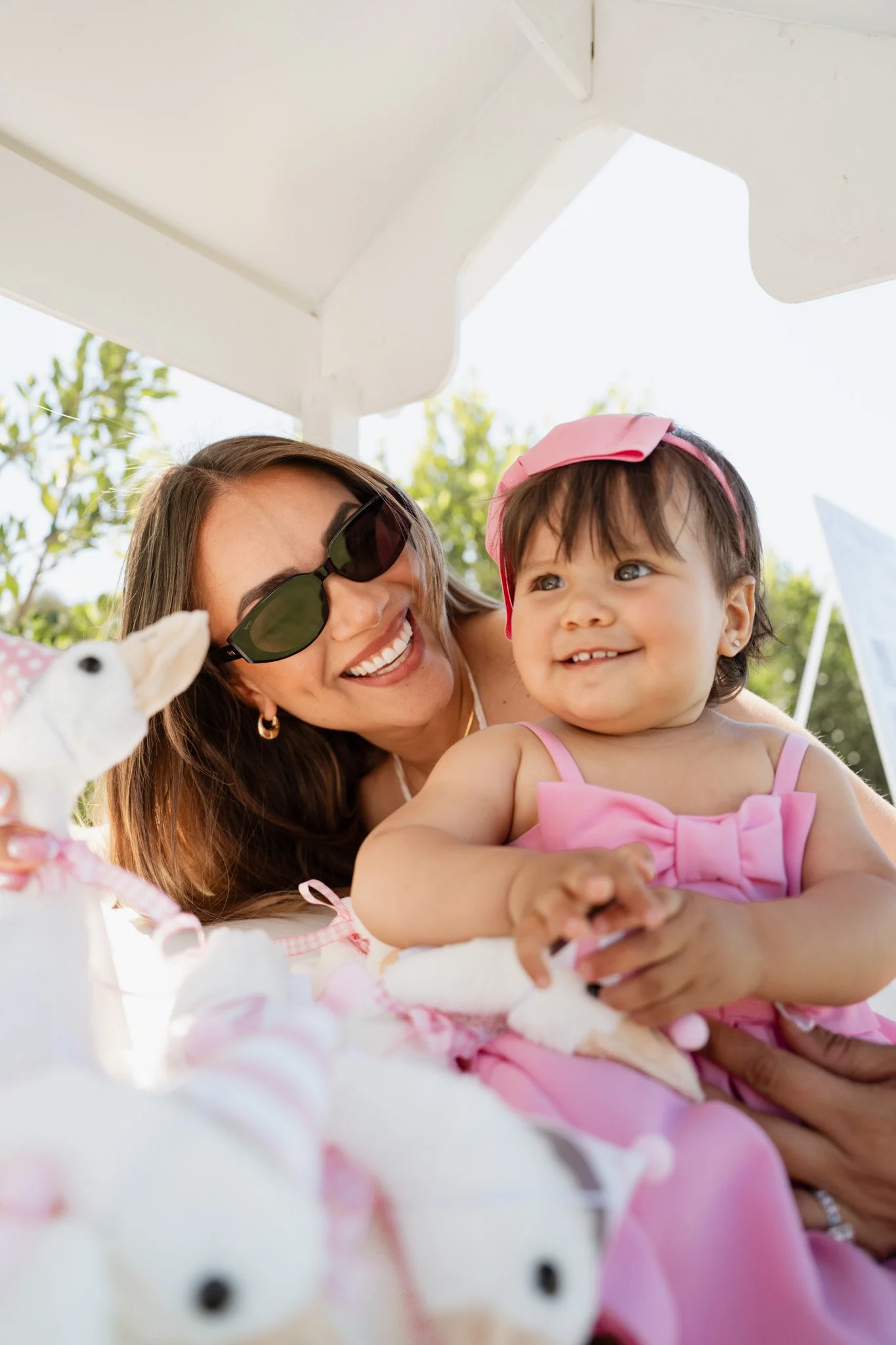 A woman wearing sunglasses and a young girl in a pink dress and pink bow smiling and playing with stuffed animals outdoors.