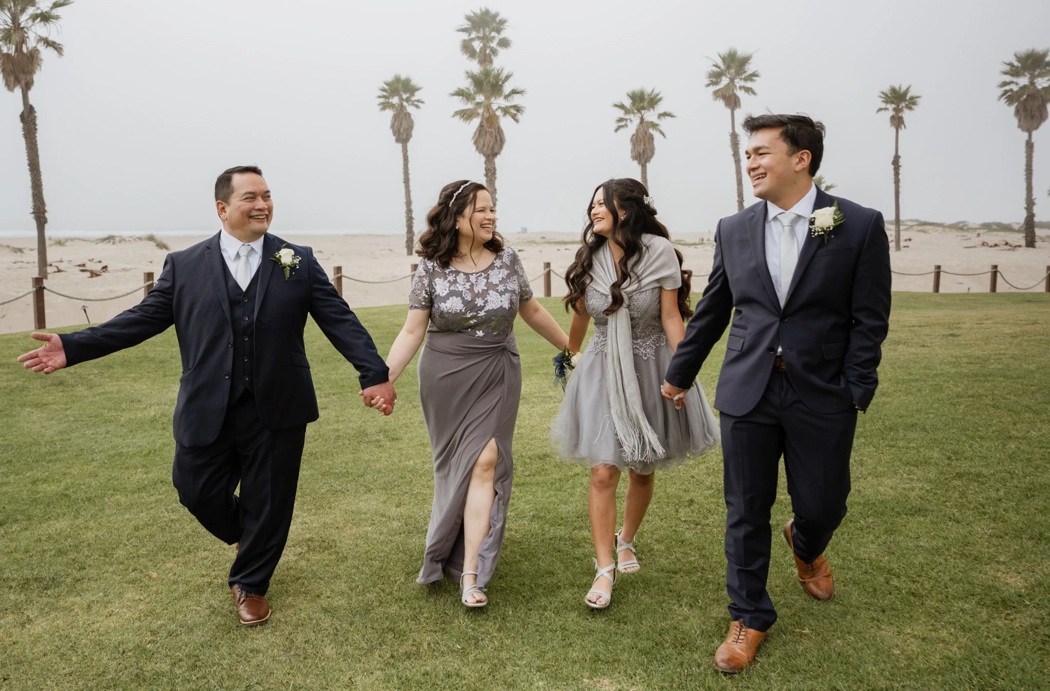 Group of four friends holding hands and walking on a grass lawn near a beach with palm trees, smiling and dressed in formal attire for a wedding celebration.