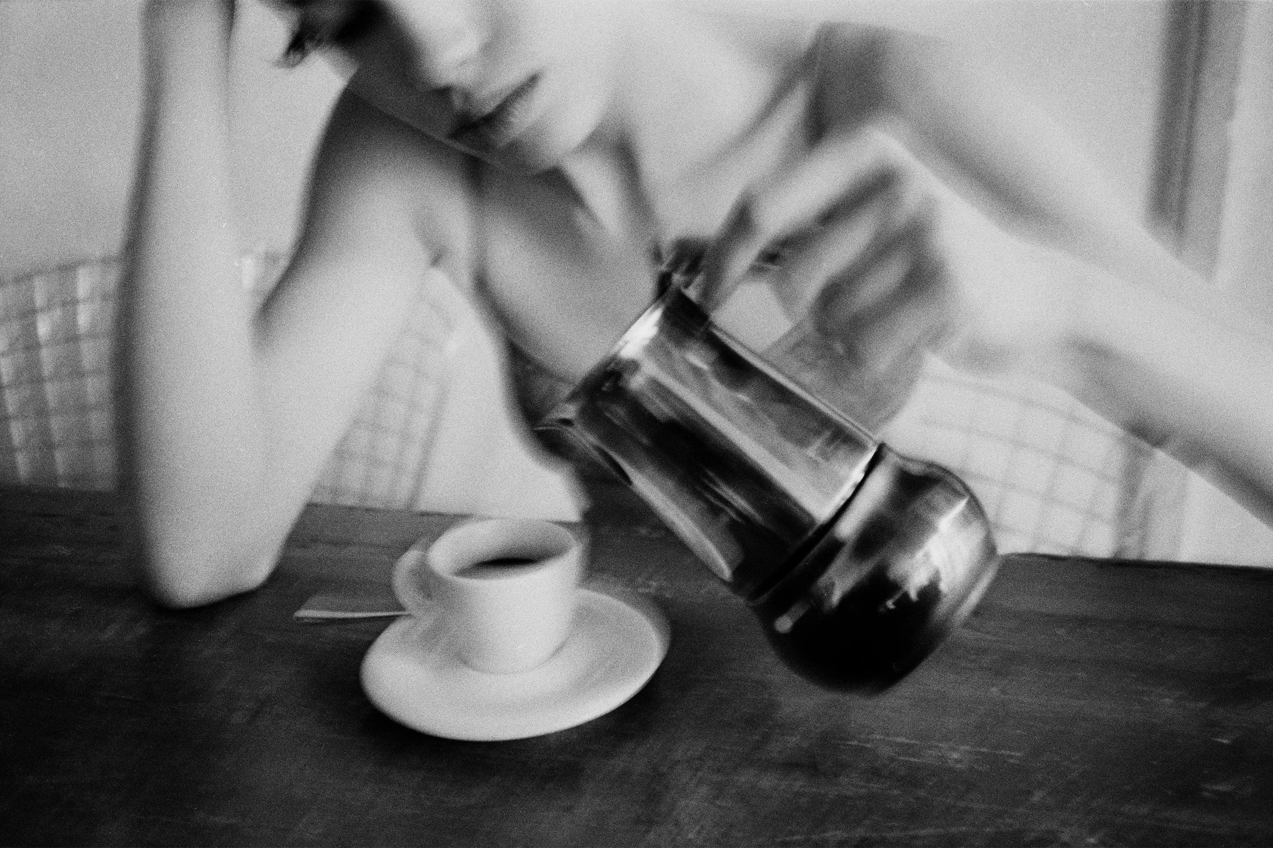 A person pouring dark coffee from a pot into a small white cup on a saucer, on a wooden table.