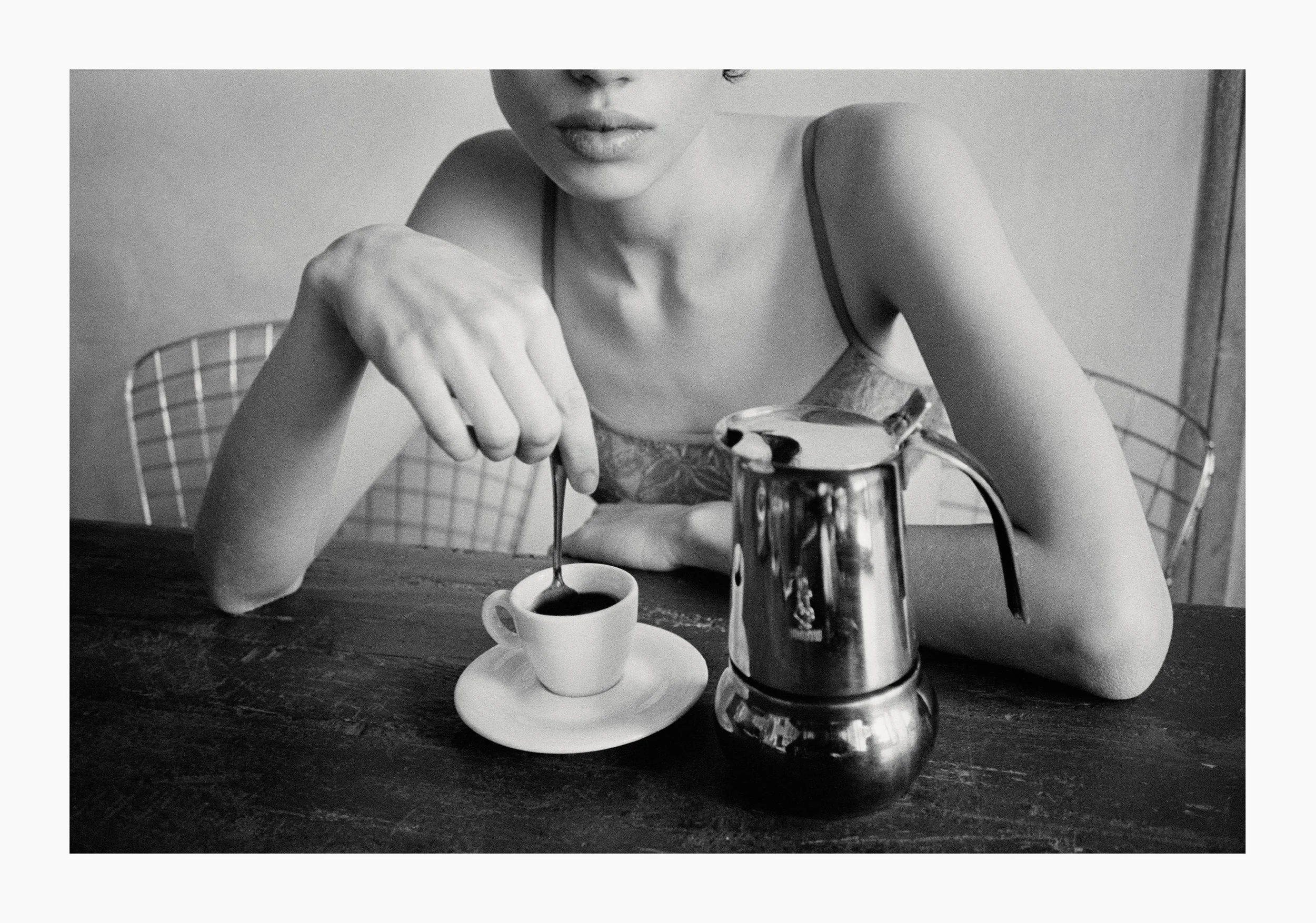 A woman in a tank top sitting at a wooden table, pouring coffee from a pot into a small cup on a saucer, with a wire chair and plain wall in background.