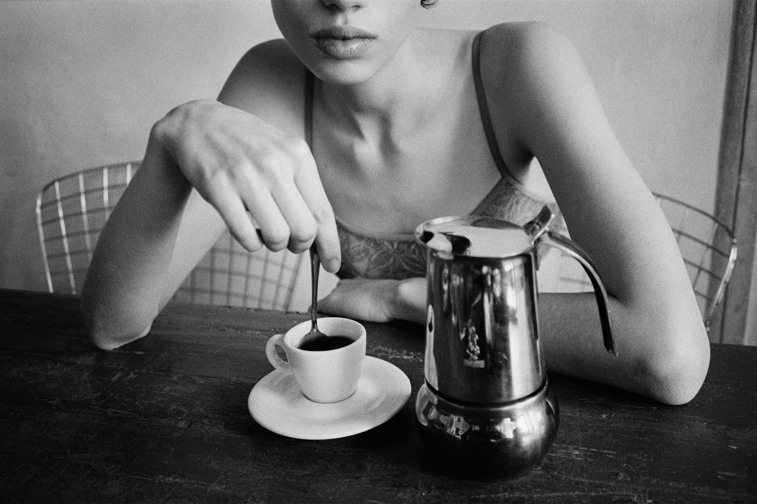 A woman with a serious expression is sitting at a wooden table and stirring a cup of coffee, with a coffee pot nearby, in a black-and-white photo.