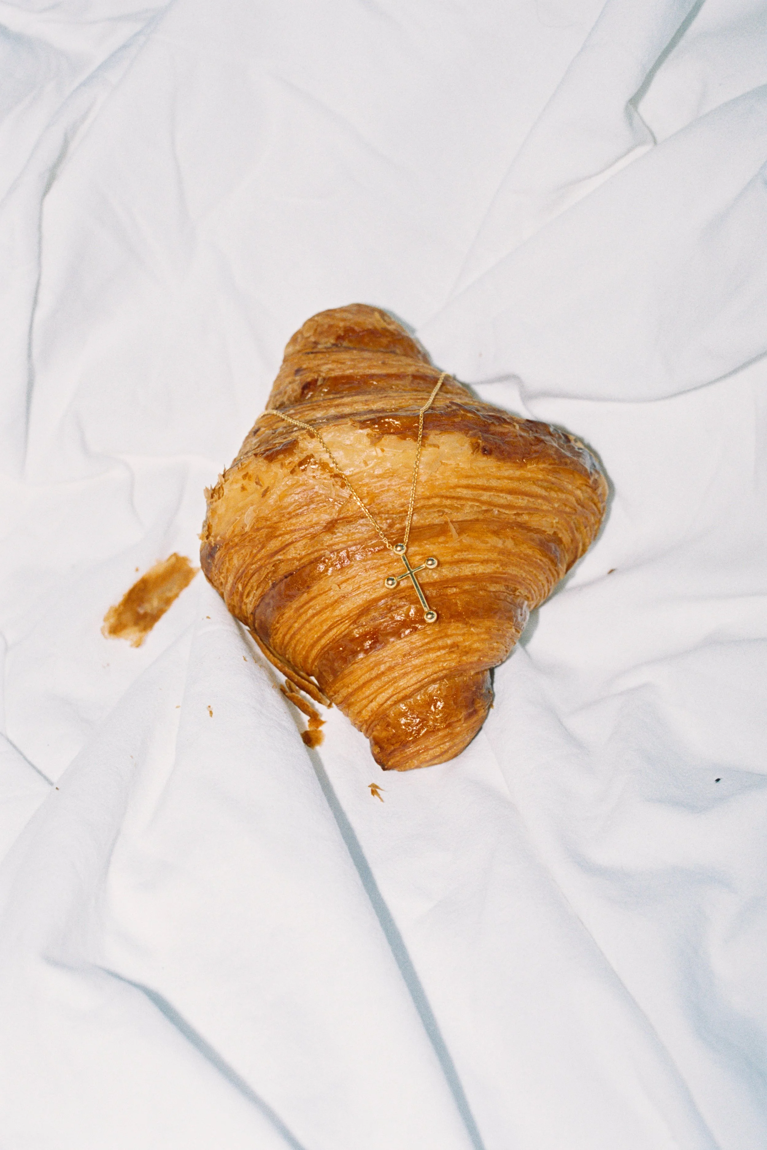 A golden brown croissant with a gold cross necklace resting on top, placed on crumpled white paper.
