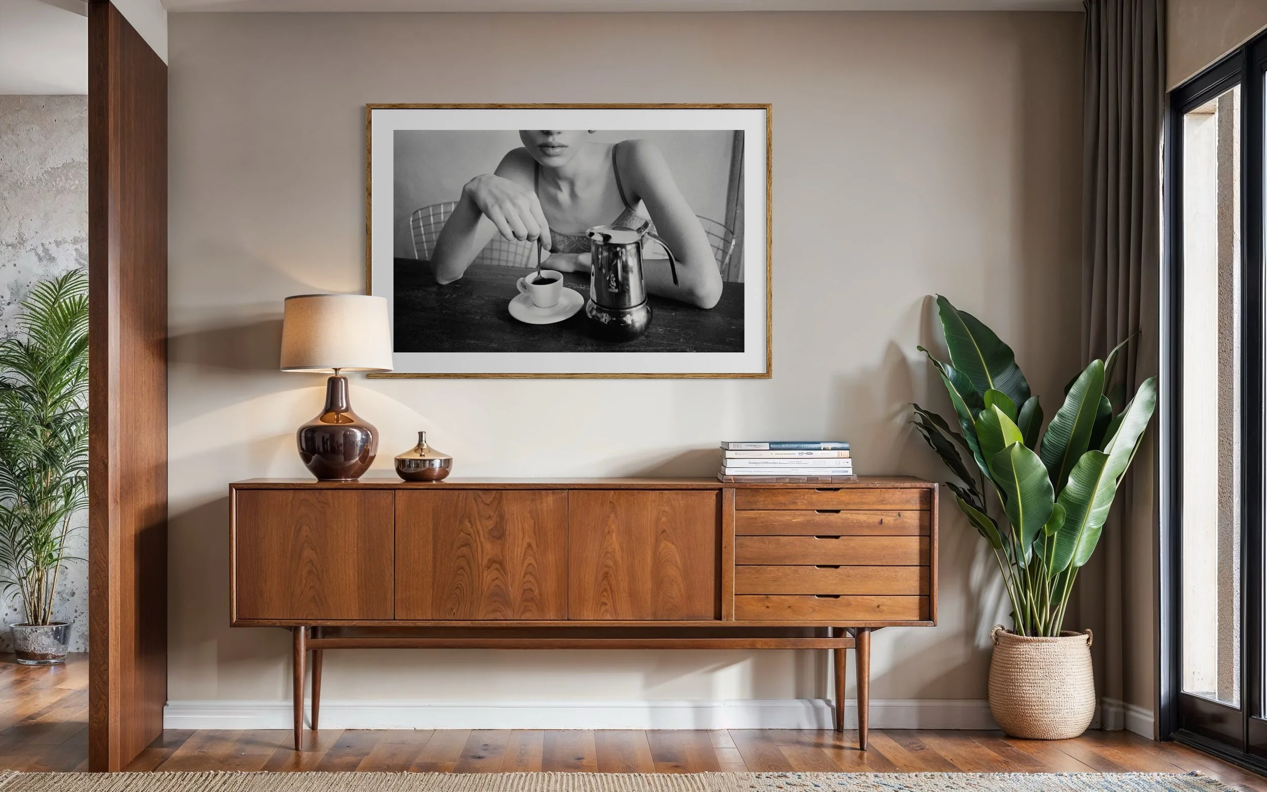 A mid-century modern wooden sideboard with a teapot, books, and a decorative item on top, next to a potted plant, and a framed black and white photograph of a woman drinking coffee hanging on the beige wall.
