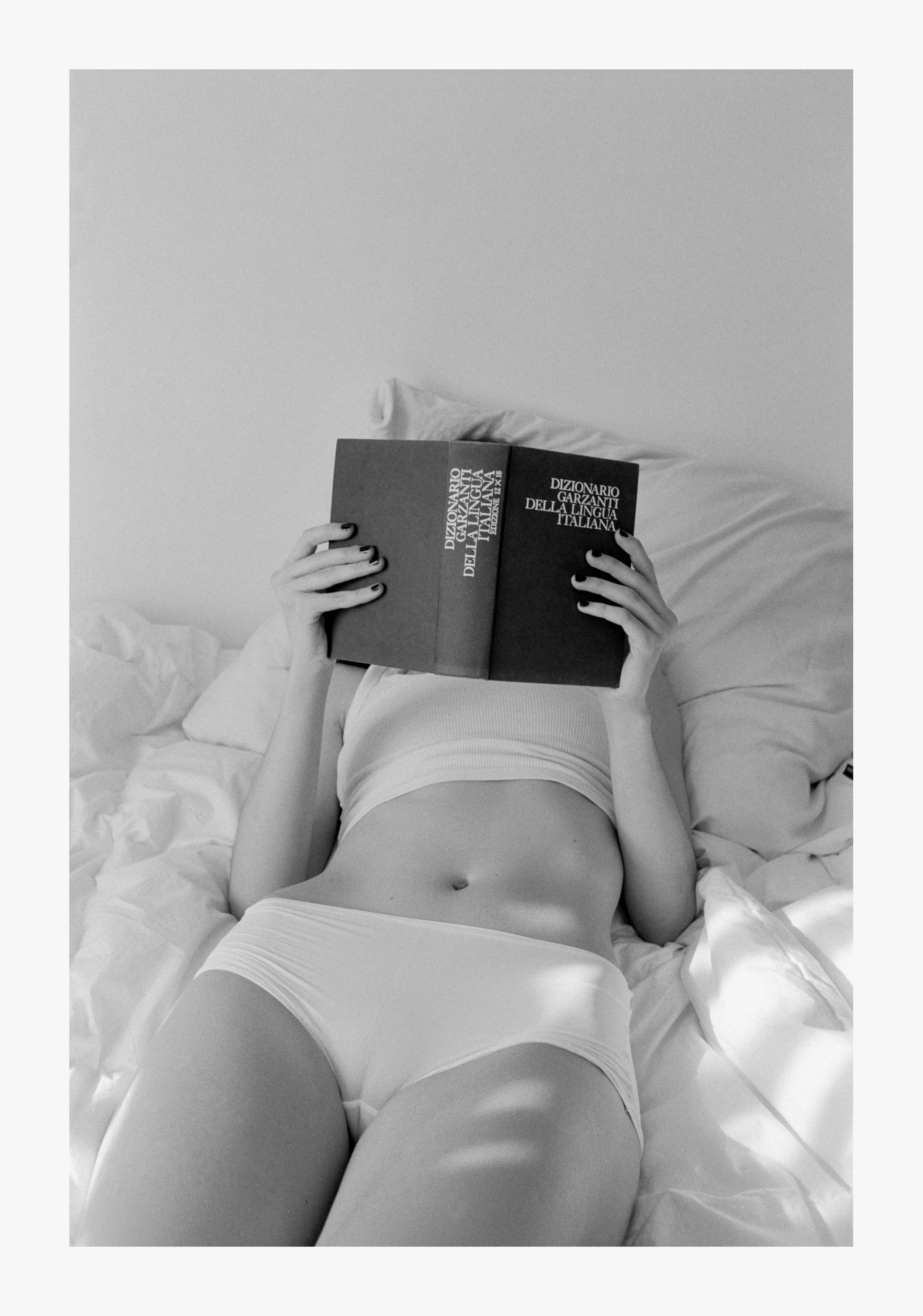 A person lying on a bed with white sheets, reading a dictionary titled "Dizionario Garzanti Della Lingua Italiana".
