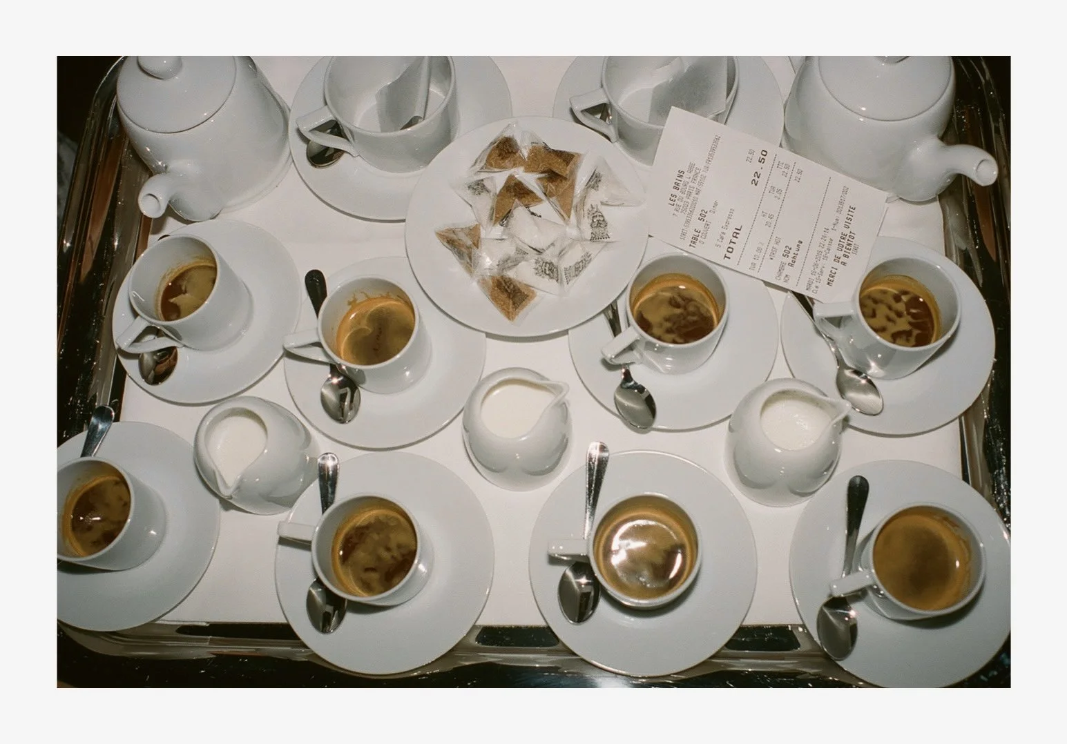 A tray with seven cups of coffee, six small pitchers of cream, one spoon, a plate with bread and an order receipt on top.