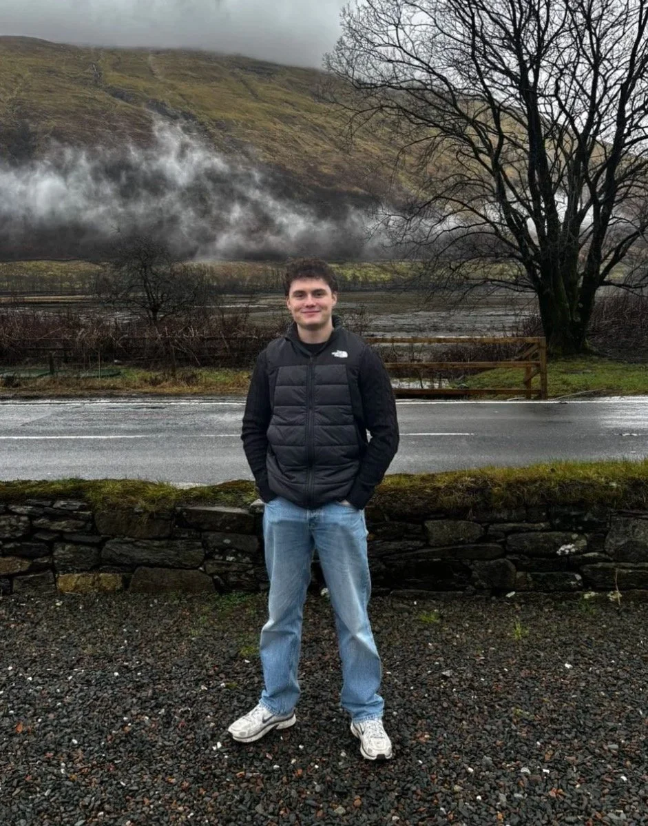 A young man standing outdoors on a gravel surface with hands in pockets, smiling, wearing a black puffer jacket, light blue jeans, and white sneakers. Behind him, there is a stone wall, a road, leafless trees, a body of water, and hills with low-hanging clouds and mist.