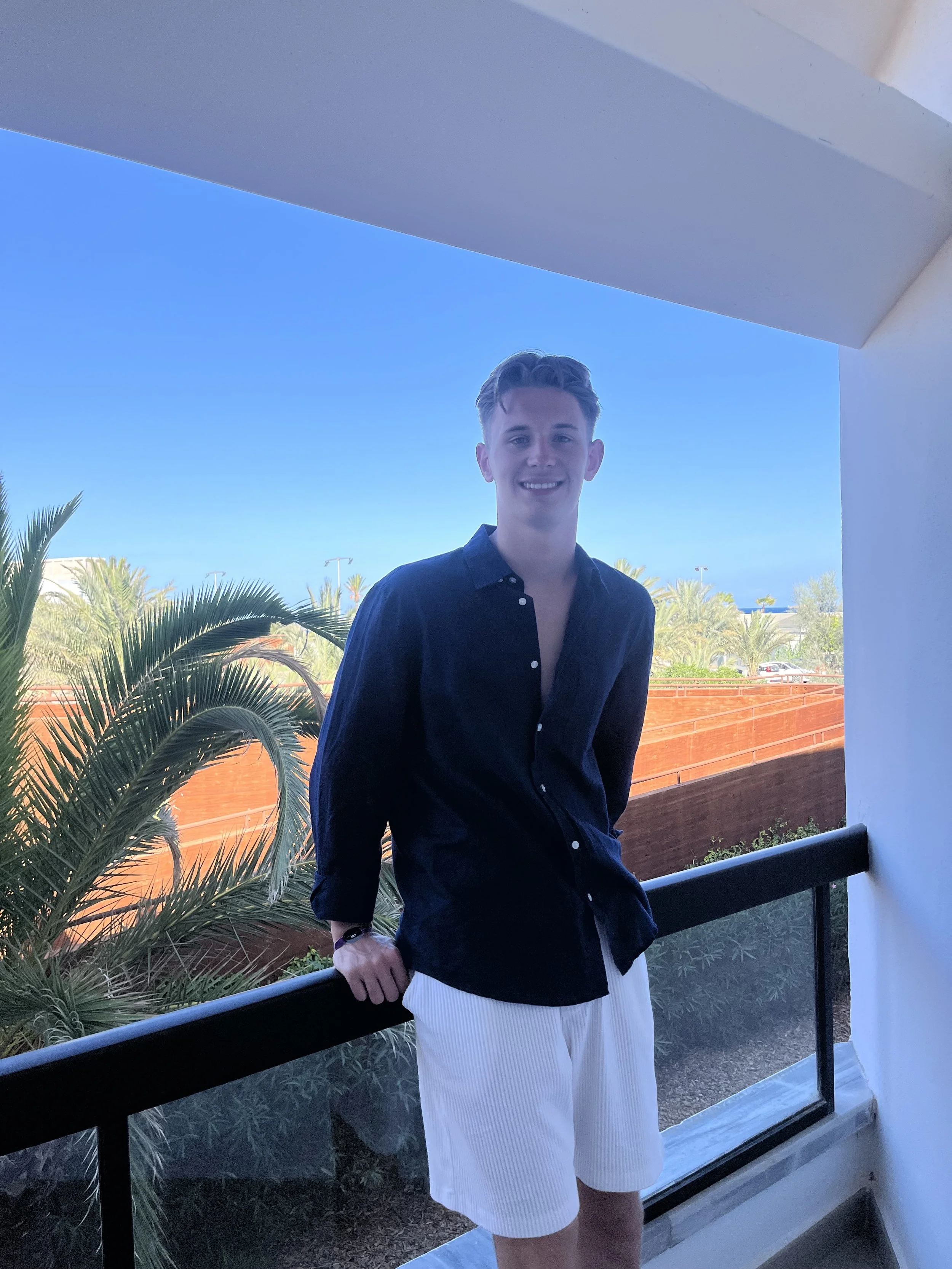 Young man smiling on balcony with palm trees and blue sky in background.