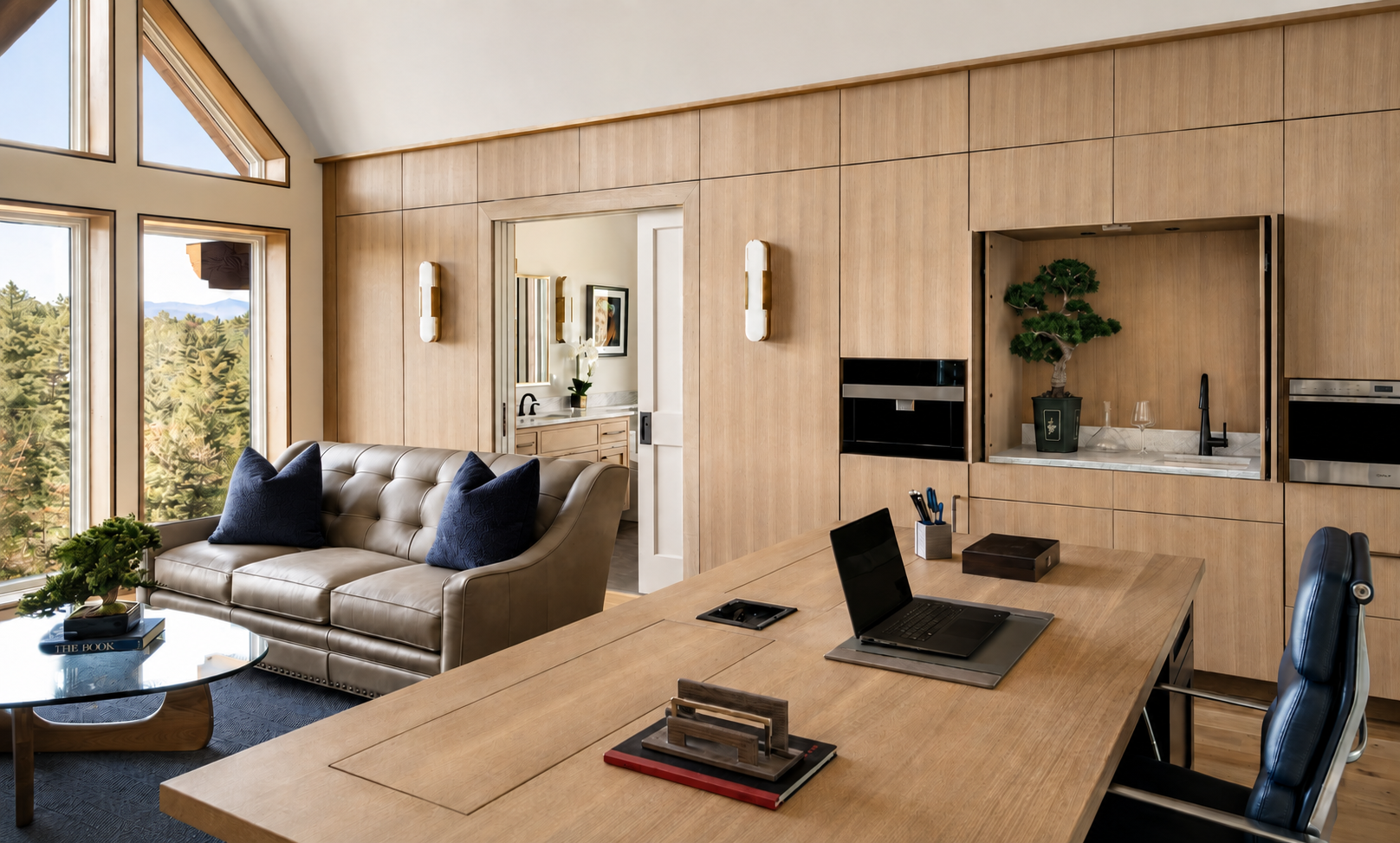 Modern living space with a beige leather sofa with blue pillows, wooden walls, a large window showing trees and mountains, and a wooden desk with a laptop and office supplies.