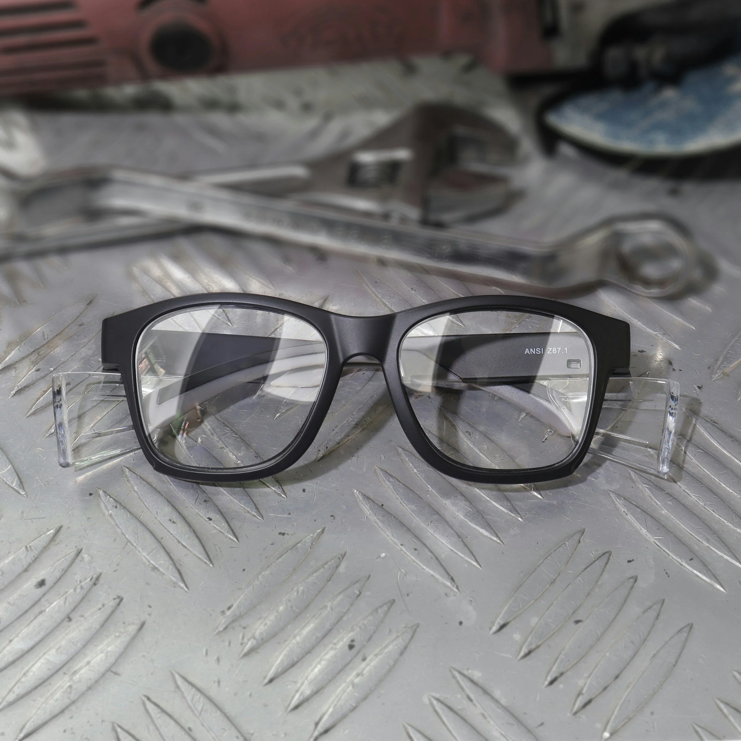 Black eyeglasses with clear plastic arms resting on a metallic, textured surface, with wrenches and a shoe in the background.