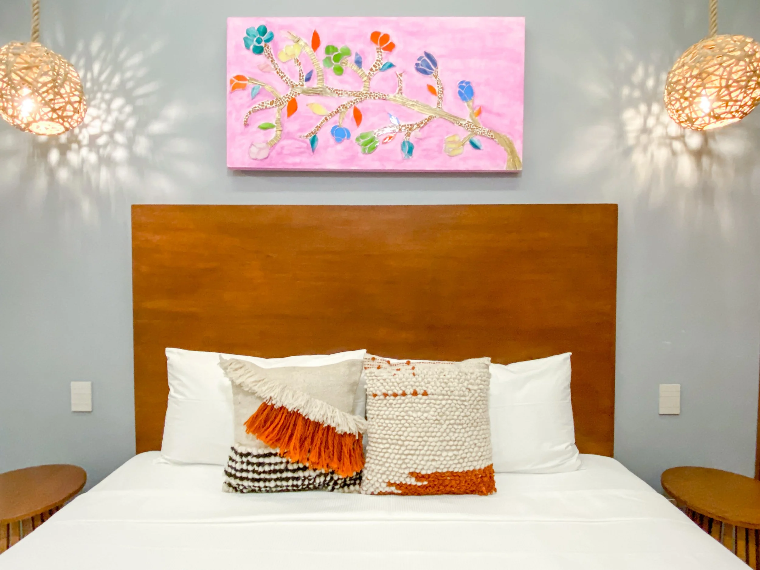 Cama con cabecera de madera, decorada con varias almohadas con texturas y colores en tonos tendencia, dos lamparas de mimbre en los lados, cuadro con rama y hojas en tonos pastel en la pared.