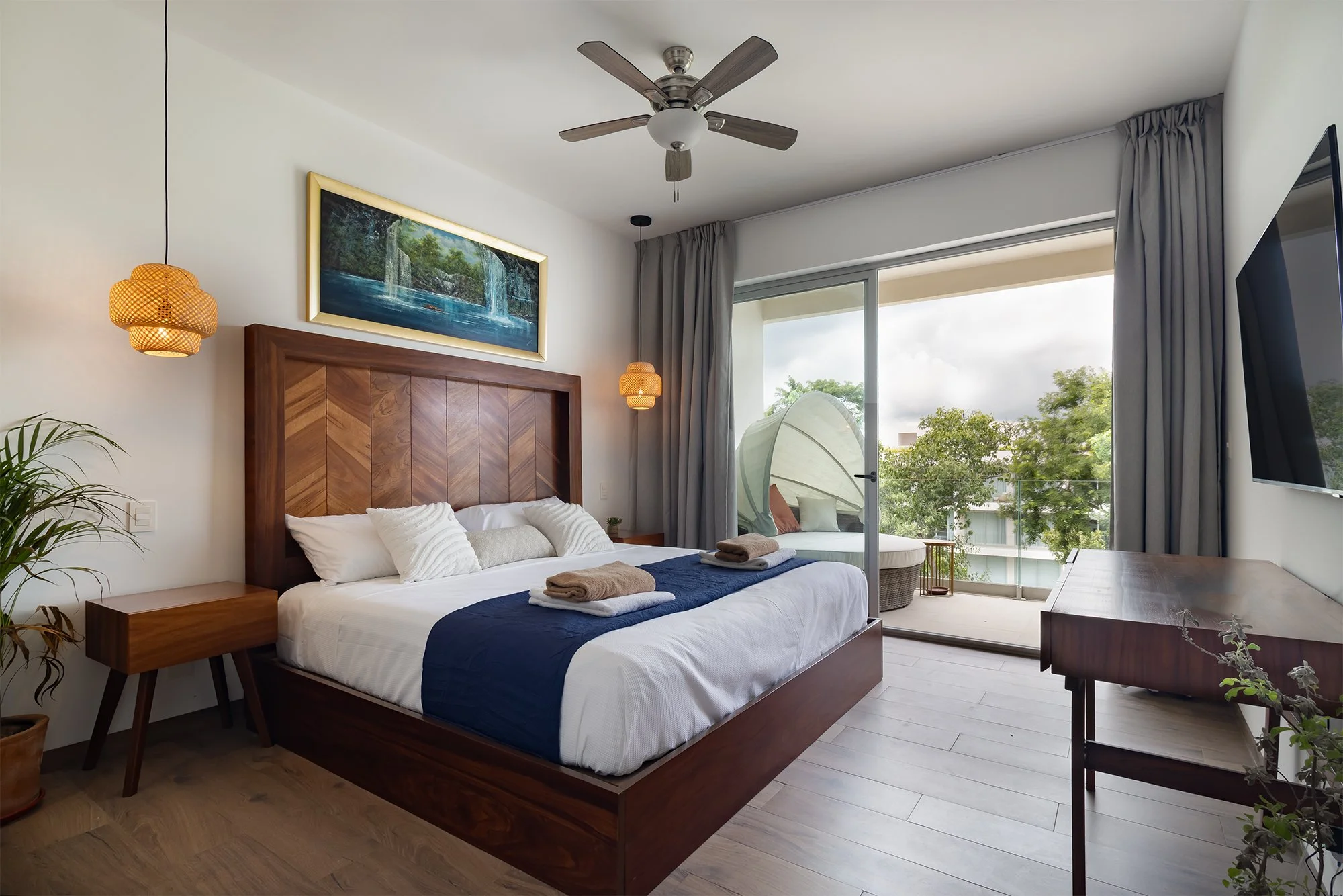 Habitación moderna con cama grande, tocador de madera, iluminación cálida, vista a un balcón con árboles, ventilador en el techo, cortinas grises, televisor grande y detalles decorativos