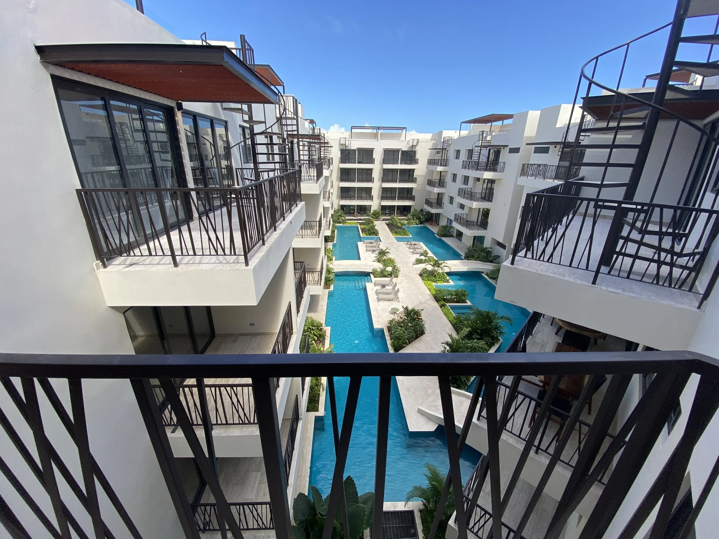 Vista desde un balcón de un complejo de apartamentos blancos con piscinas y áreas verdes, bajo un cielo claro y azul.