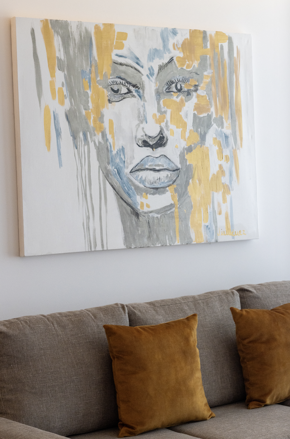 Cuadro de pintura abstracta de rostro de mujer con detalles en gris, azul y amarillo, montado en la pared sobre un sofá de tapizado gris y cojines marrones.