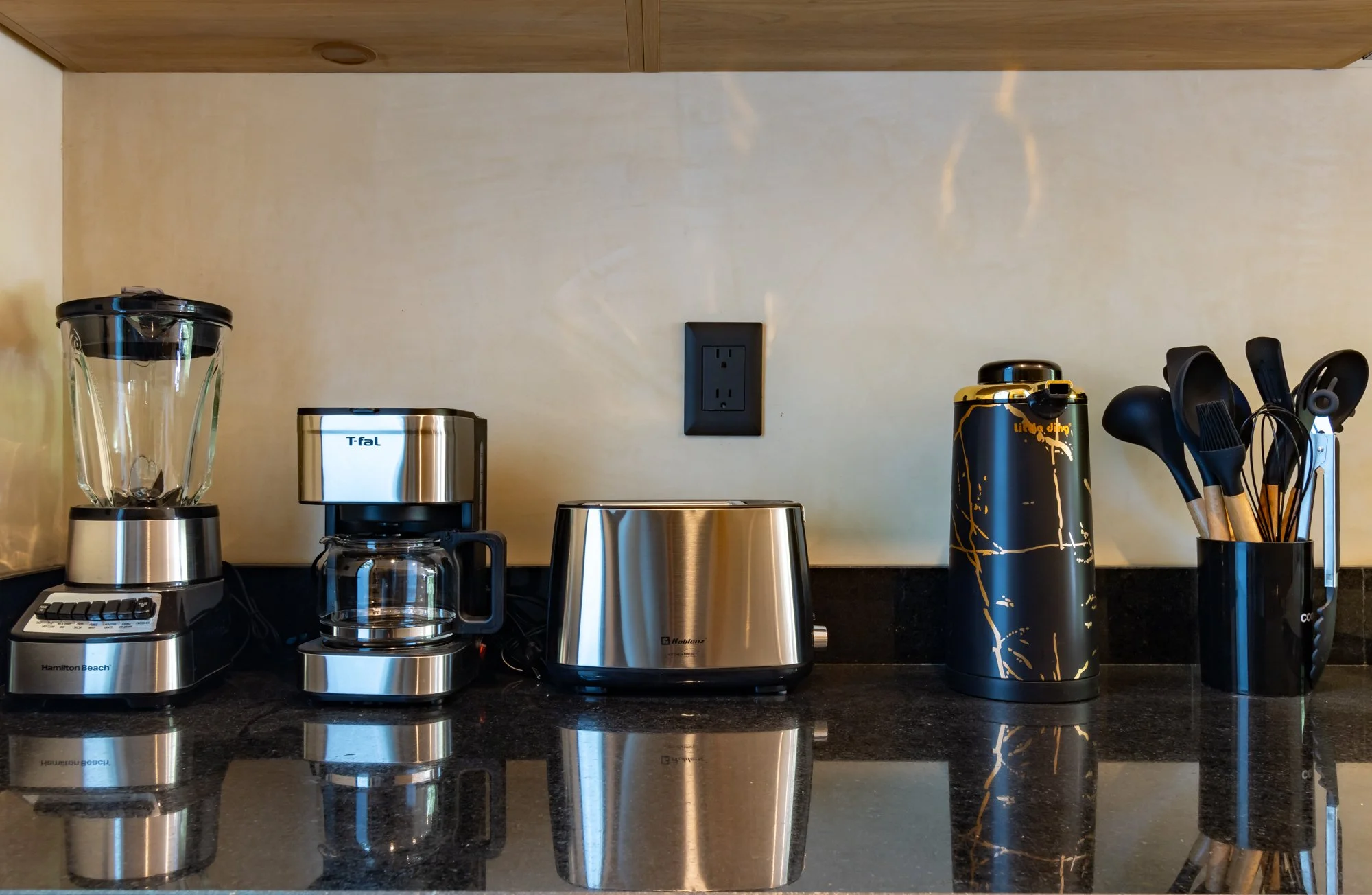 Estantes con objetos de cocina incluyendo procesador de alimentos, cafetera, termo, y utensilios de cocina, sobre una encimera de piedra negra.