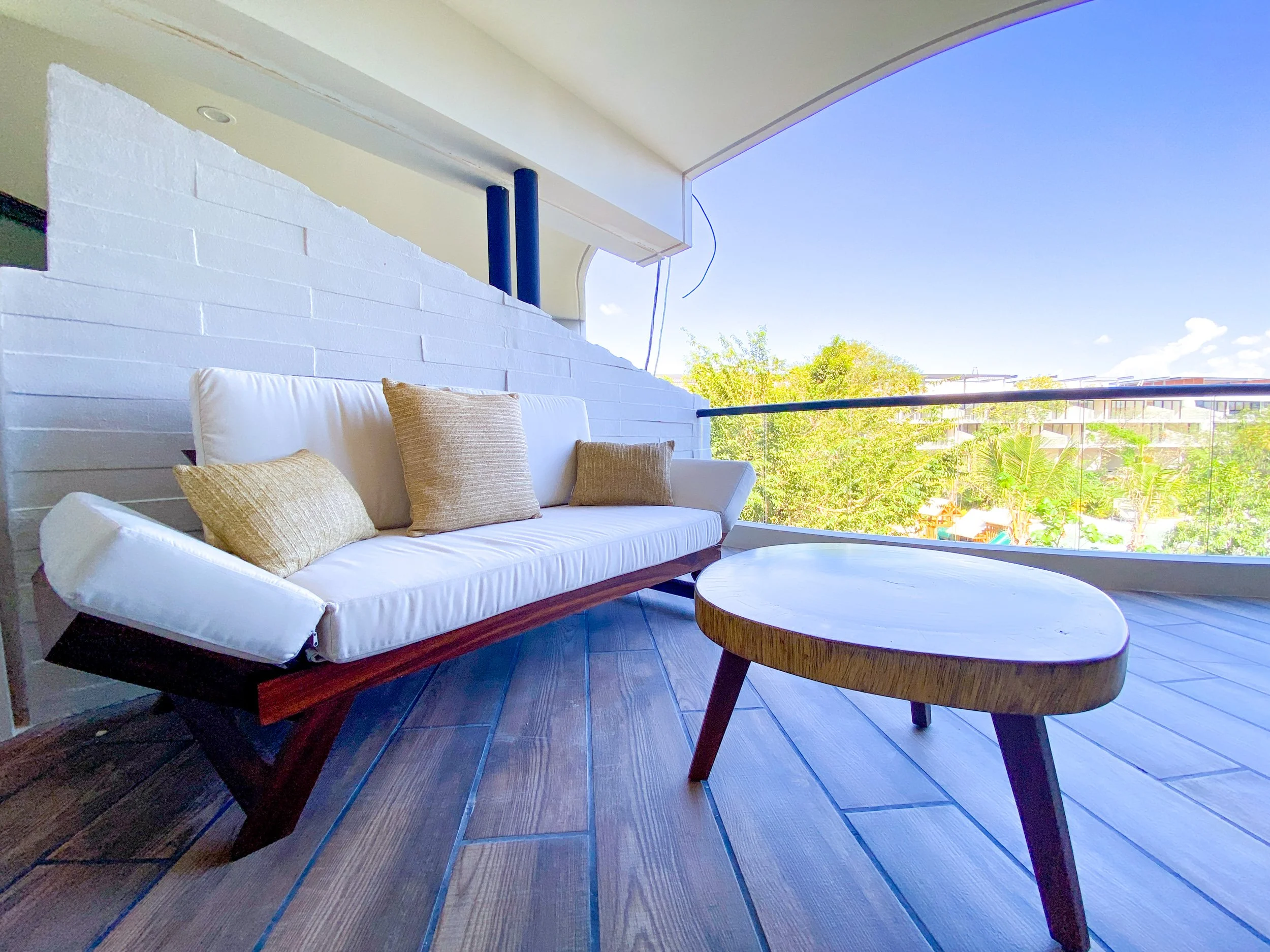 Un balcón con sillones y mesas, con vista a árboles y edificios en un día soleado y despejado.