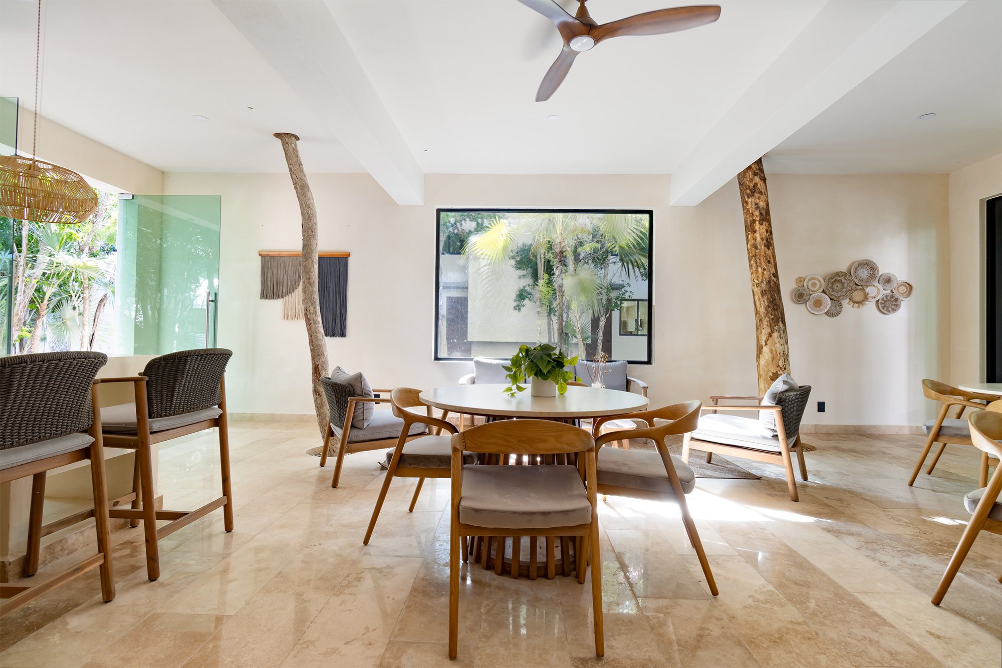 Sala de estar con sillones de madera y cojines, mesas redondas, árboles en el interior, ventana grande con vista a plantas exteriores, decoraciones de paredes y techo con ventilador de madera.