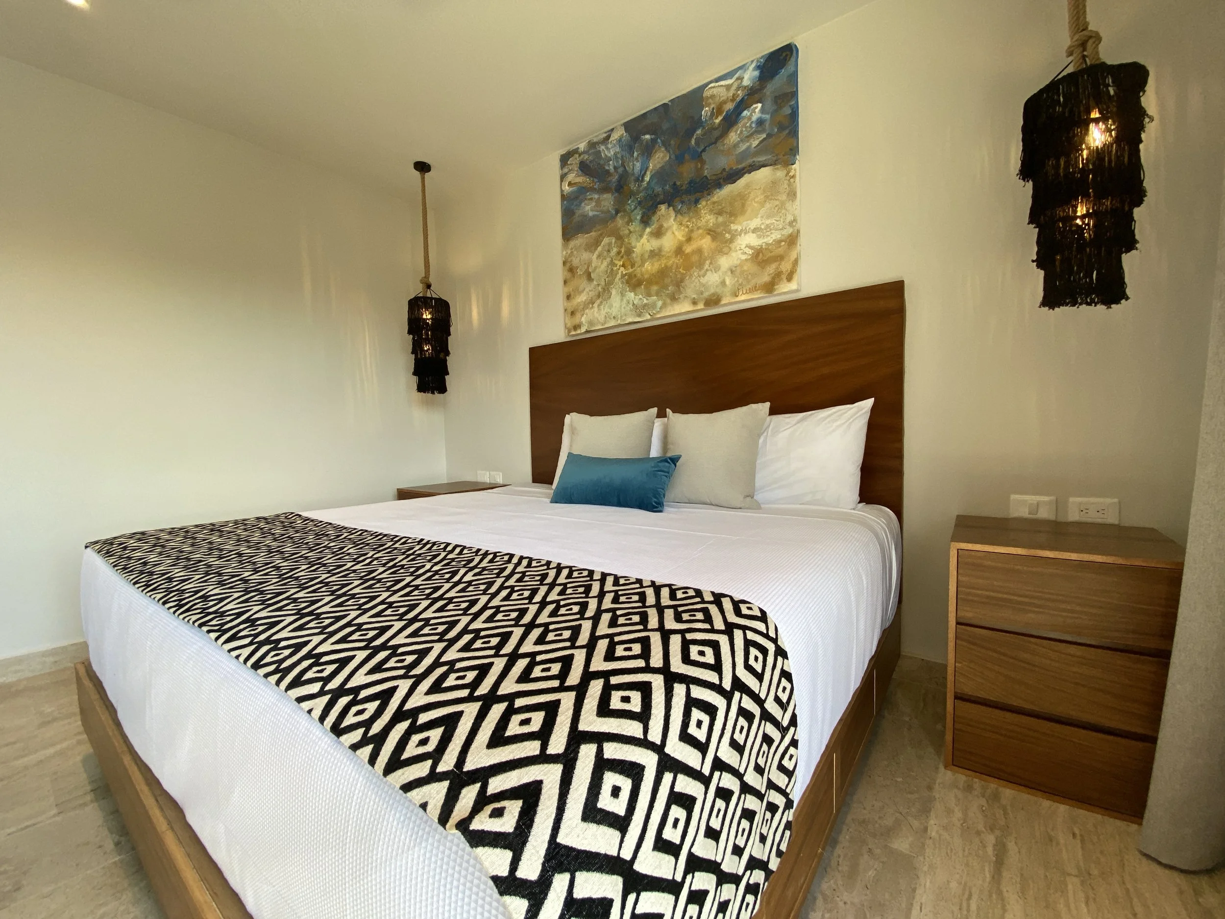 Cama con cabecera de madera, dos almohadas blancas y una almohada azul, lámparas colgantes de fibra negra a cada lado, cuadro abstracto en la pared, noche con techo blanco y suelo de madera.