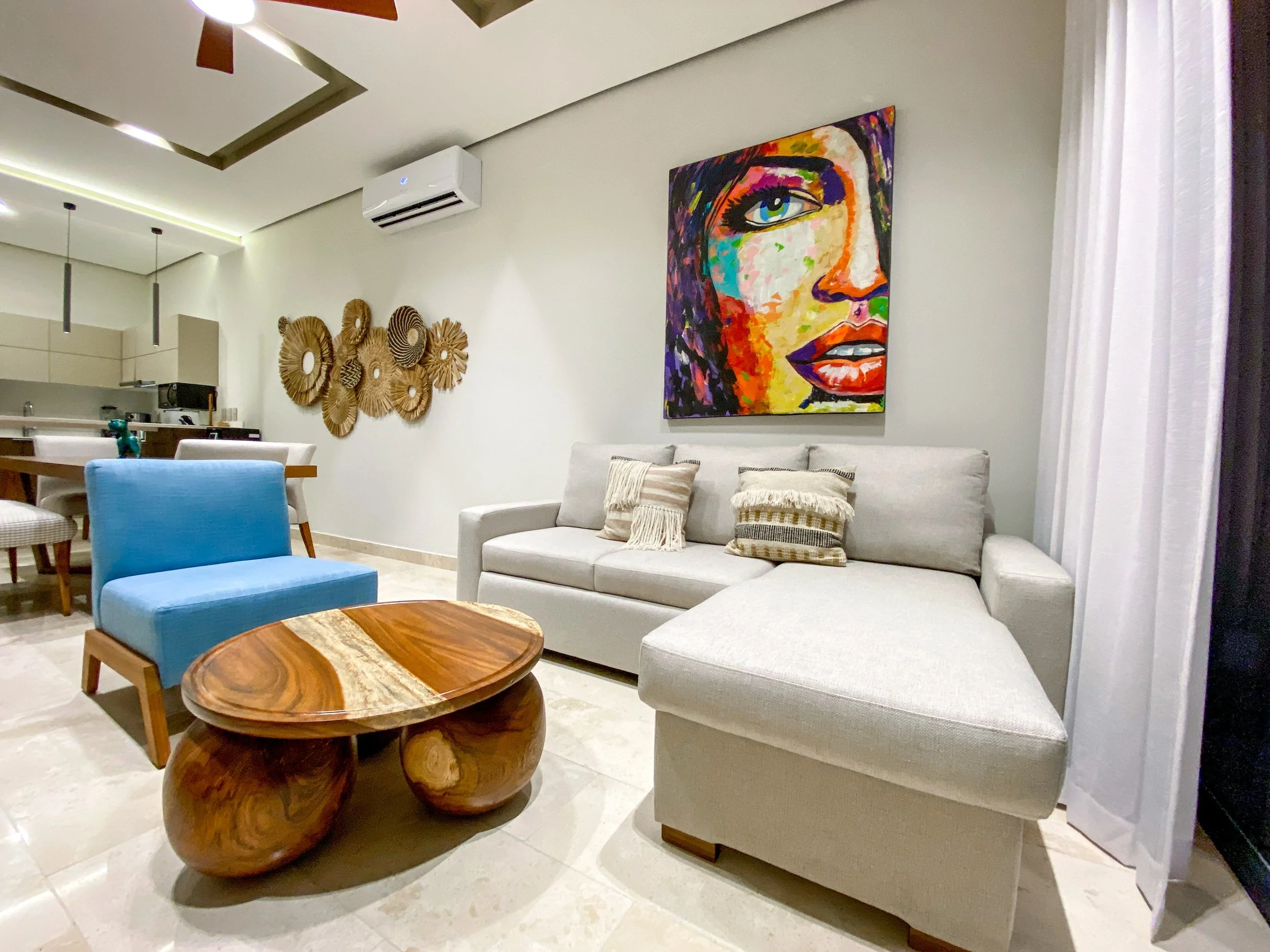 Sala de estar con sofá gris, cojines, mesa de madera, obra de arte colorida de rostro femenino en la pared, decoraciones de madera en la pared, cortinas blancas, aire acondicionado en la pared.