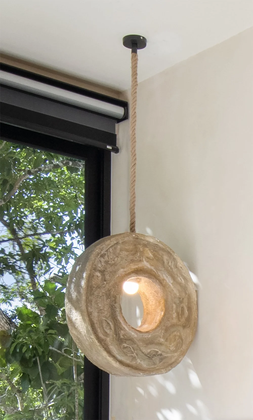 Lámpara colgante de diseño rústico hecha de piedra con agujero central, suspendida del techo por una cuerda de fibra natural, frente a una ventana con vista a árboles y hojas verdes.