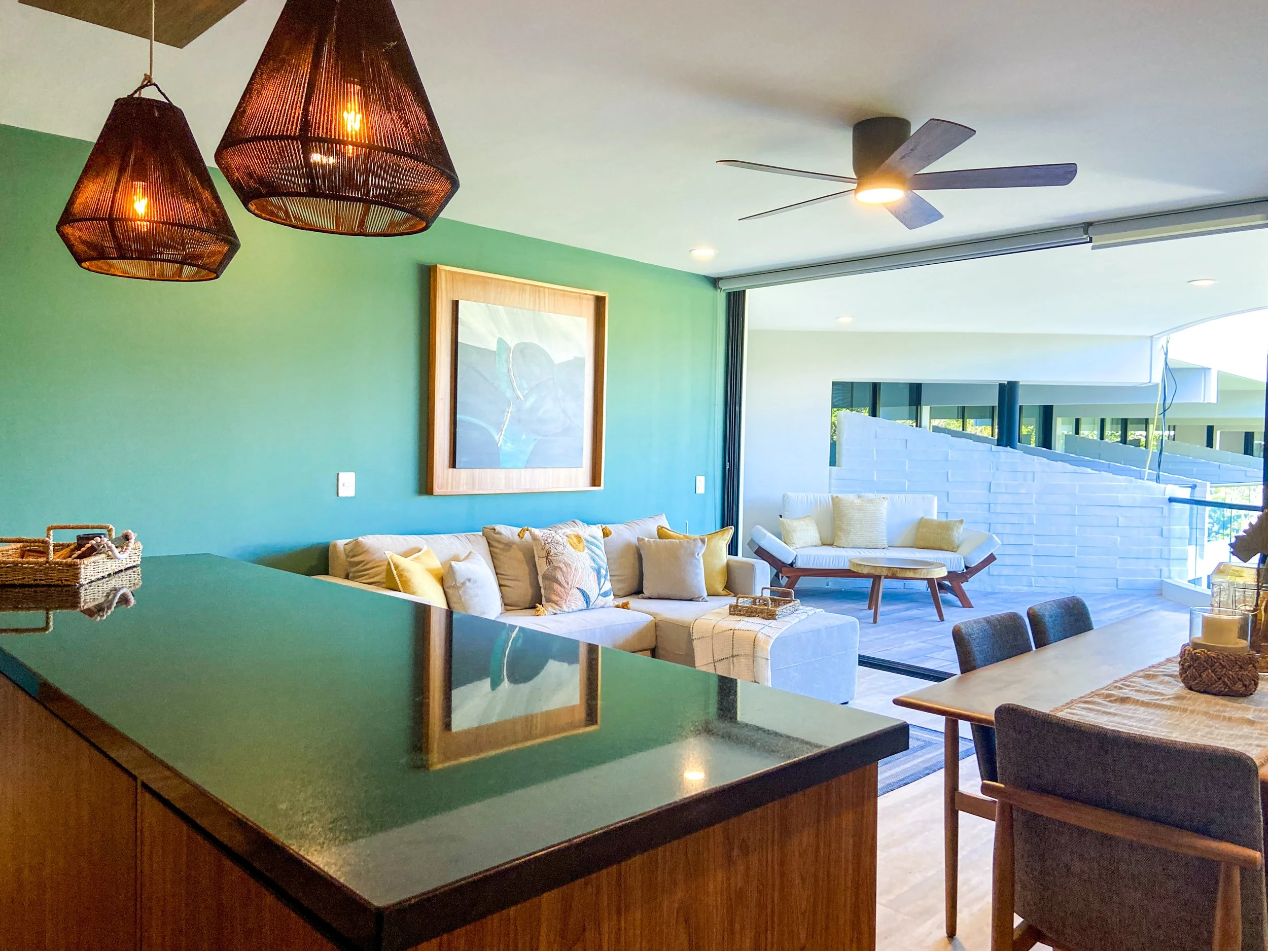 Vista de sala y comedor en una vivienda moderna con paredes verdes, un sillón con cojines, lámparas colgantes de mimbre y ventilador en el techo, y una gran ventana que permite entrada de luz natural.